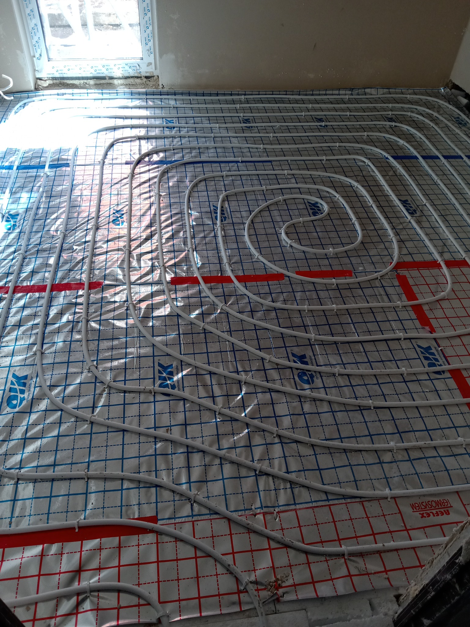 Białe rurki ogrzewania podłogowego ułożone w spiralny wzór na folii izolacyjnej z siatką, widoczne okno w tle.