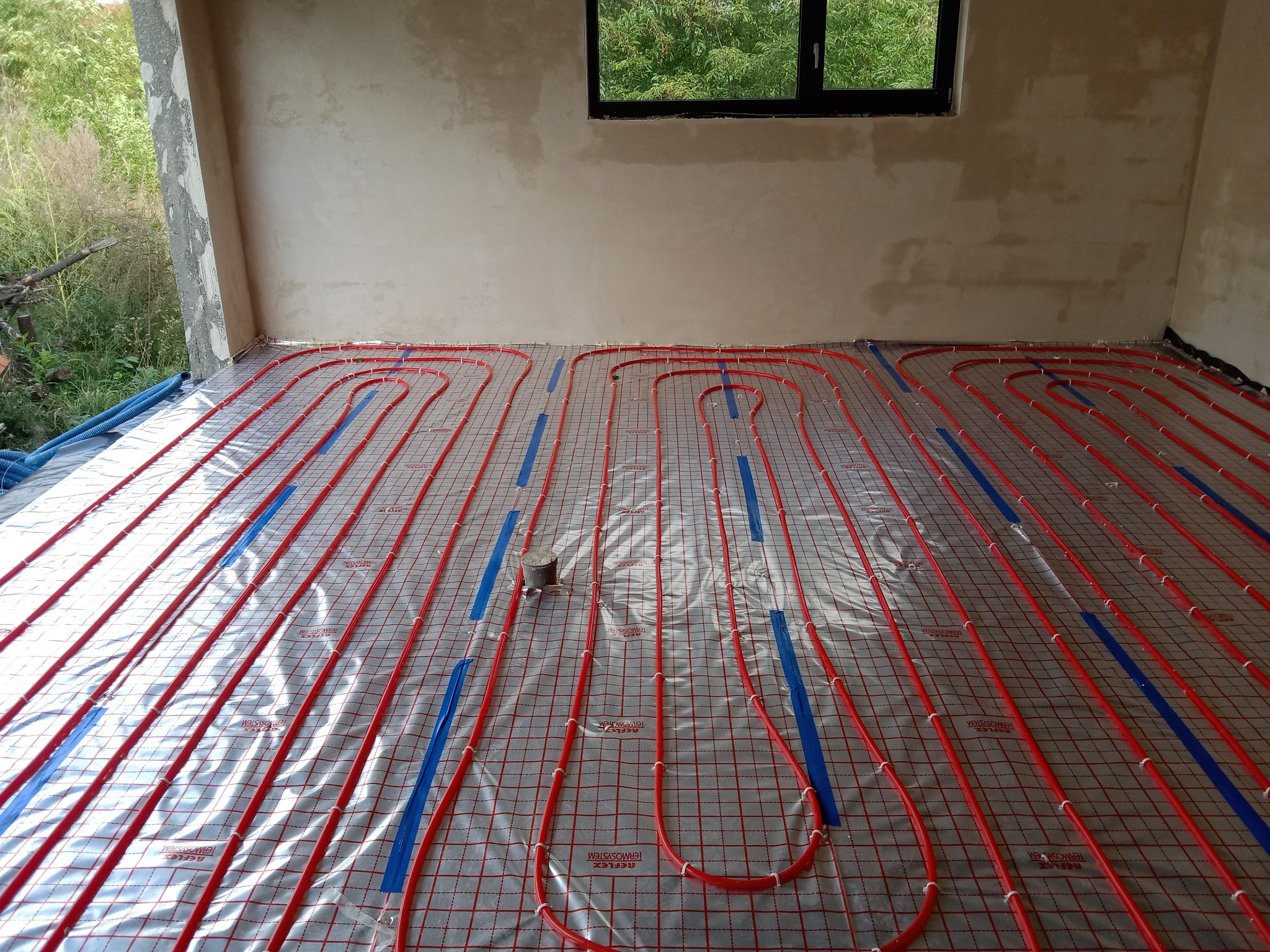 Rozłożona instalacja ogrzewania podłogowego z czerwonymi rurkami na srebrnej folii z czerwoną kratką, przytwierdzona niebieską taśmą, widok z góry w surowym wnętrzu z oknem.