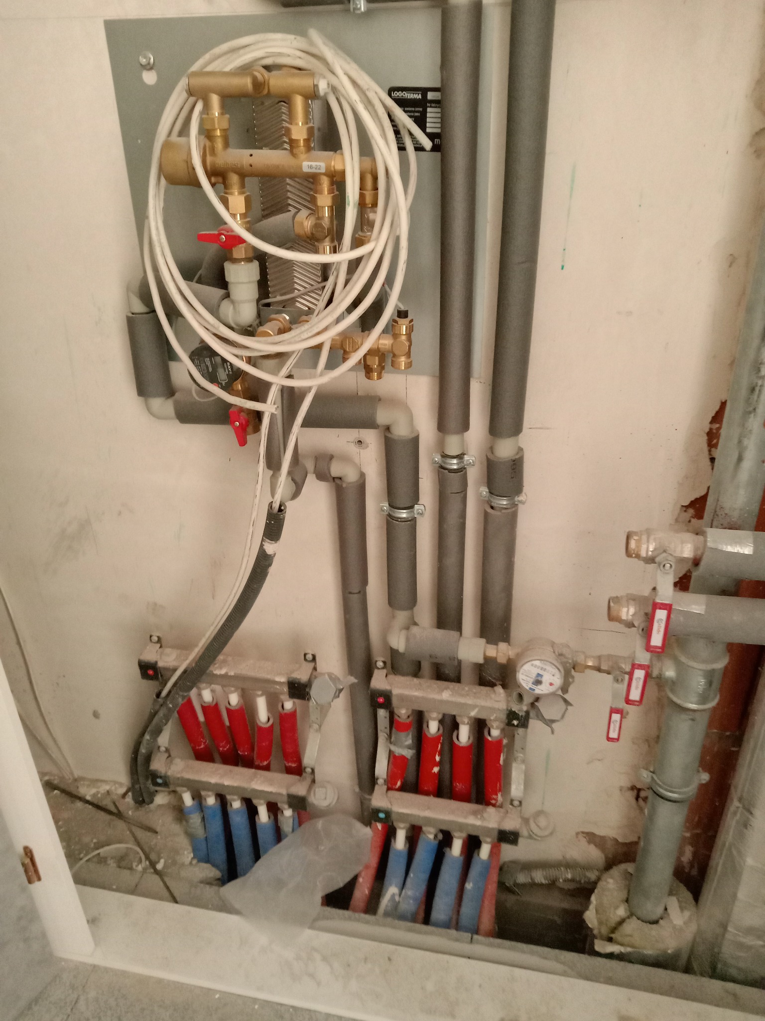 Rozdzielacz ogrzewania podłogowego z czerwono-niebieskimi pętlami, rury z izolacją termiczną, zawory i licznik na tle surowej ściany w trakcie remontu.