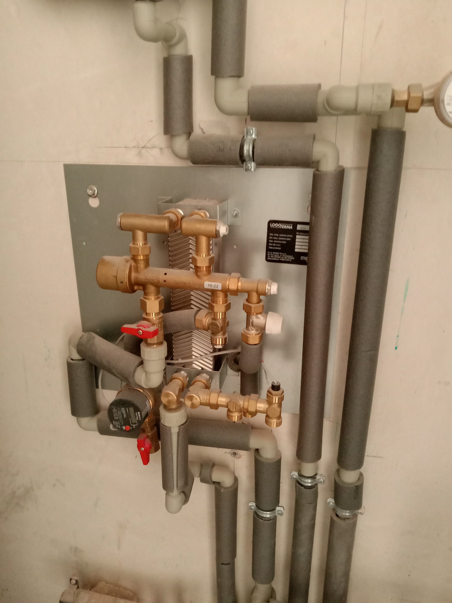 Węzeł cieplny z pompą obiegową, zaworami i rurami zaizolowanymi szarą pianką, zamontowany na metalowej płycie na tle surowej, białej ściany.