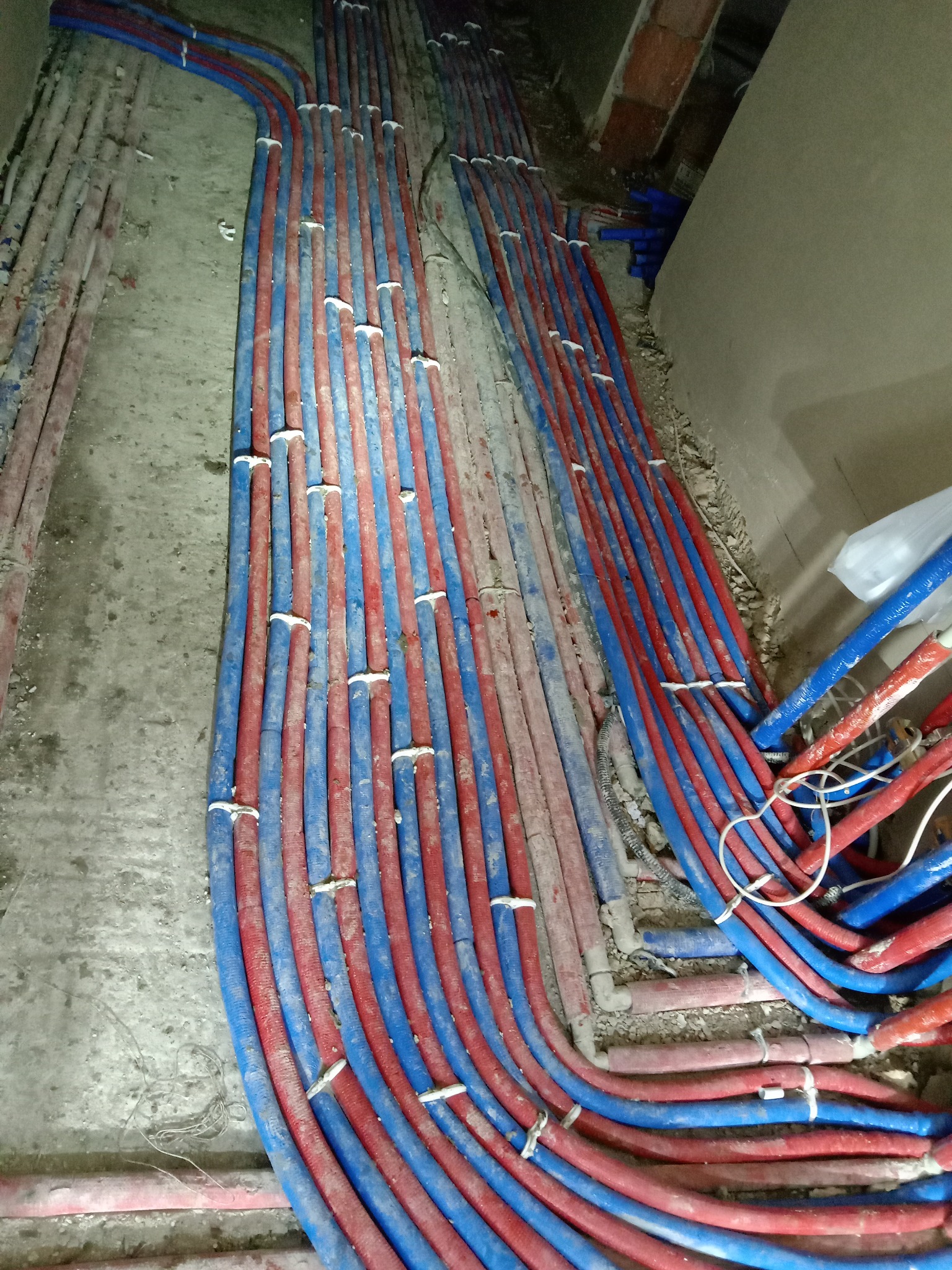 Równolegle ułożone, niebiesko-czerwone rury instalacji ogrzewania podłogowego, częściowo zalane w szarym betonie, z widocznymi białymi opaskami mocującymi.