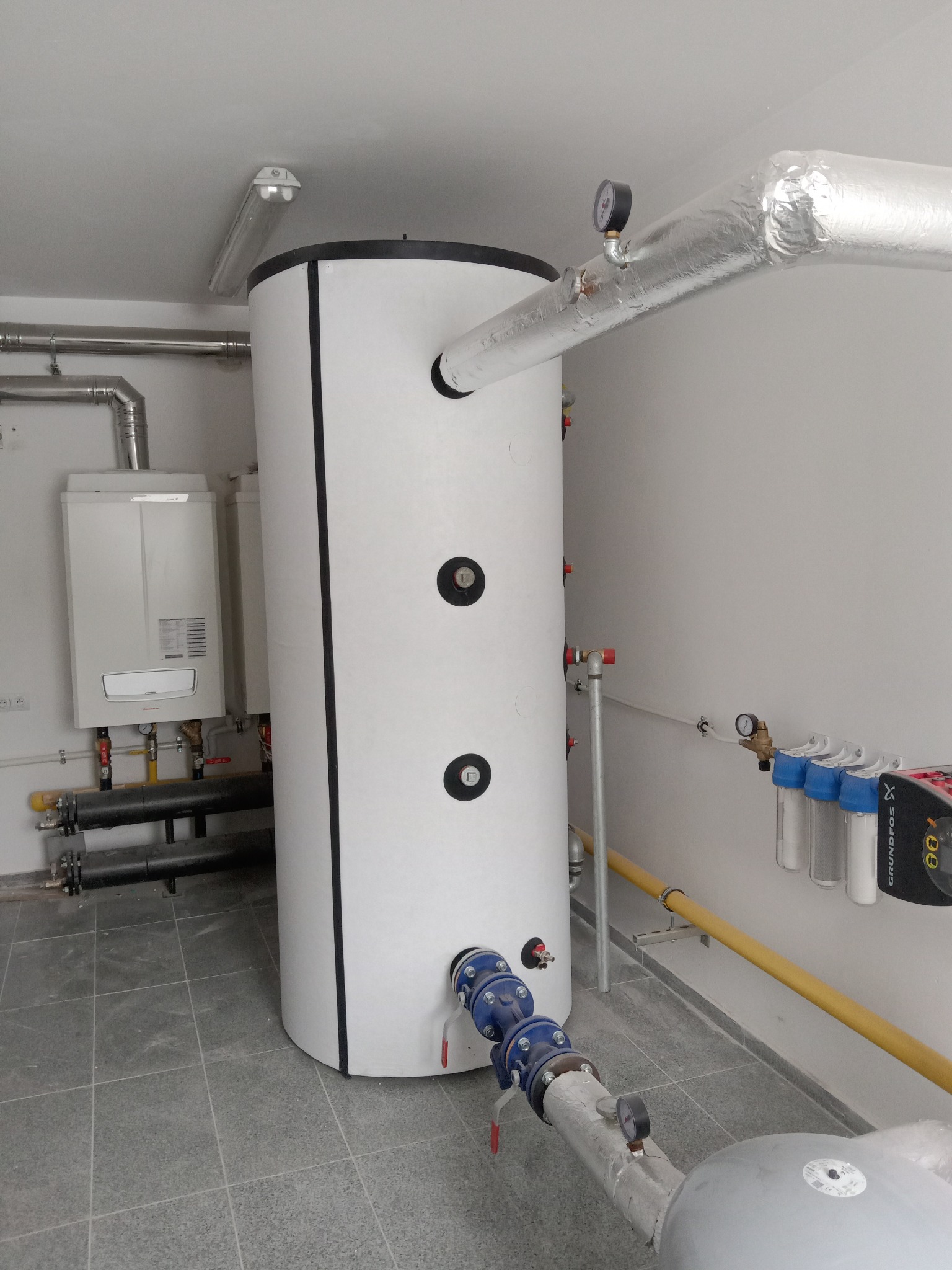Biały, cylindryczny zbiornik akumulacyjny z czarnymi okrągłymi zaślepkami, podłączony do instalacji, obok wiszący piec gazowy i filtry wody, w pomieszczeniu technicznym z szarą podłogą.