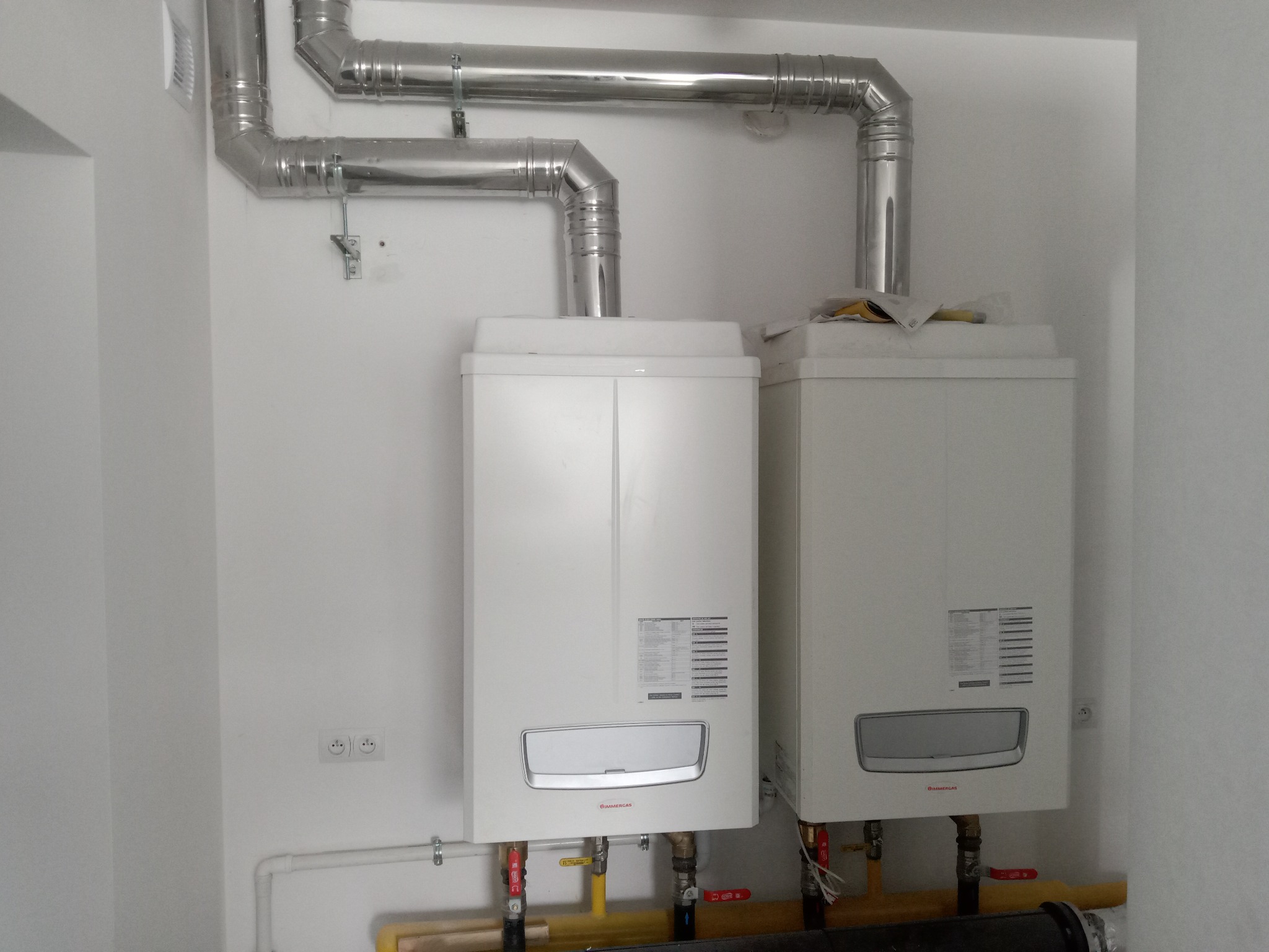 Dwa białe, wiszące kotły gazowe marki Immergas z podłączonymi rurami odprowadzającymi spaliny, zamontowane na białej ścianie w nowym pomieszczeniu z widocznymi gniazdkami elektrycznymi i rurami...