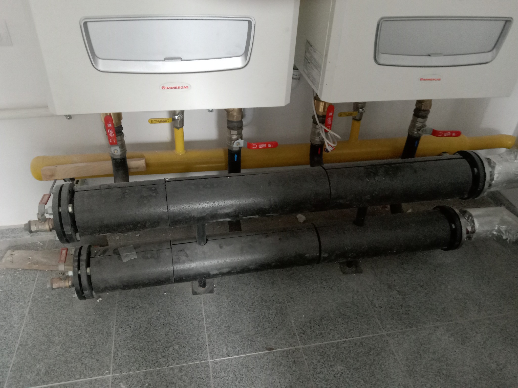 Dwa białe kotły gazowe marki Immergas, zamontowane na ścianie, z widocznymi żółtymi rurami gazowymi i czarnymi rurami instalacji grzewczej z zaworami, na tle szarej podłogi z płytek.