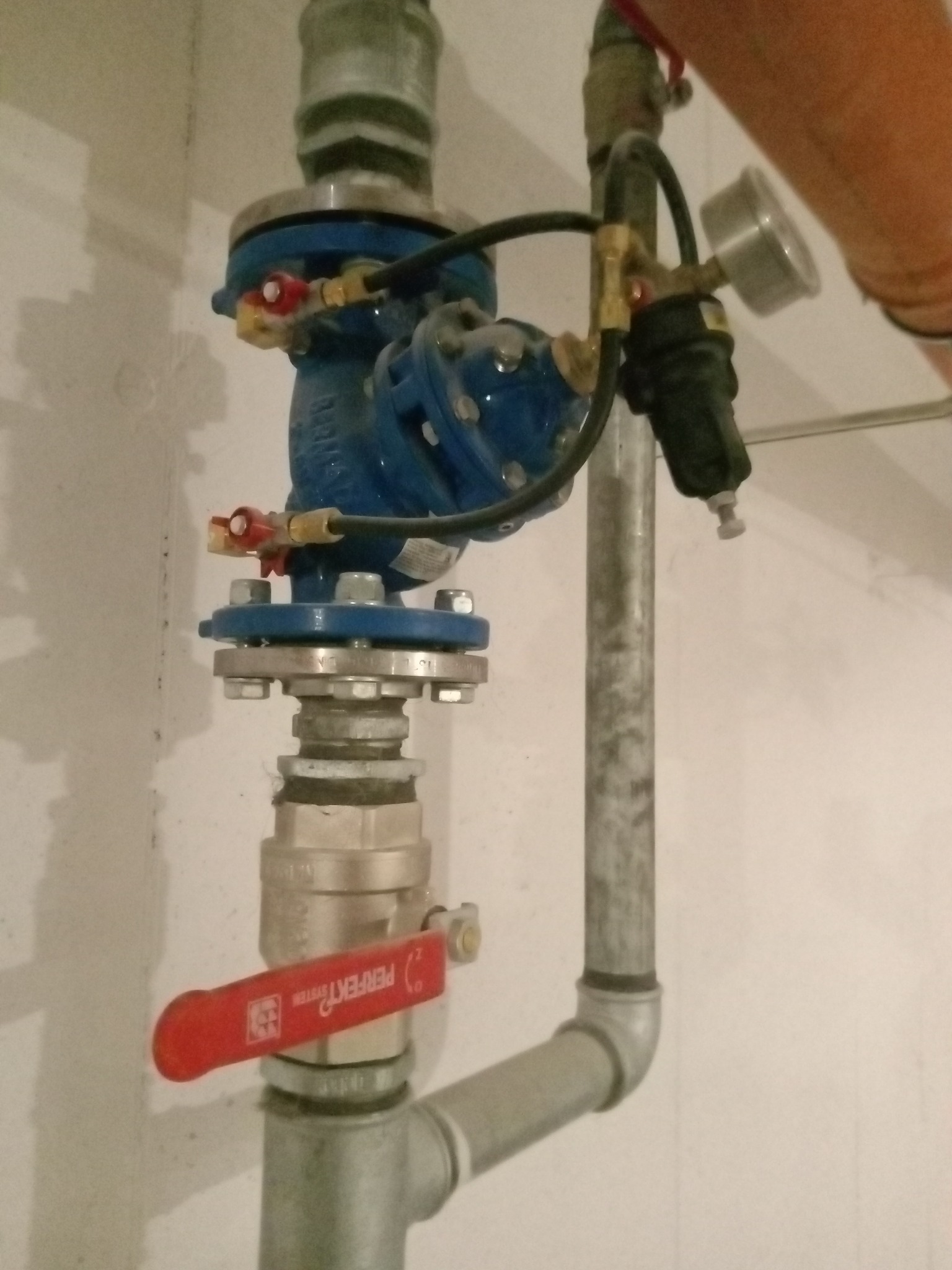 Niebieski zawór z manometrem i rurami w instalacji wodnej, widoczny czerwony uchwyt zaworu Perfect System.
