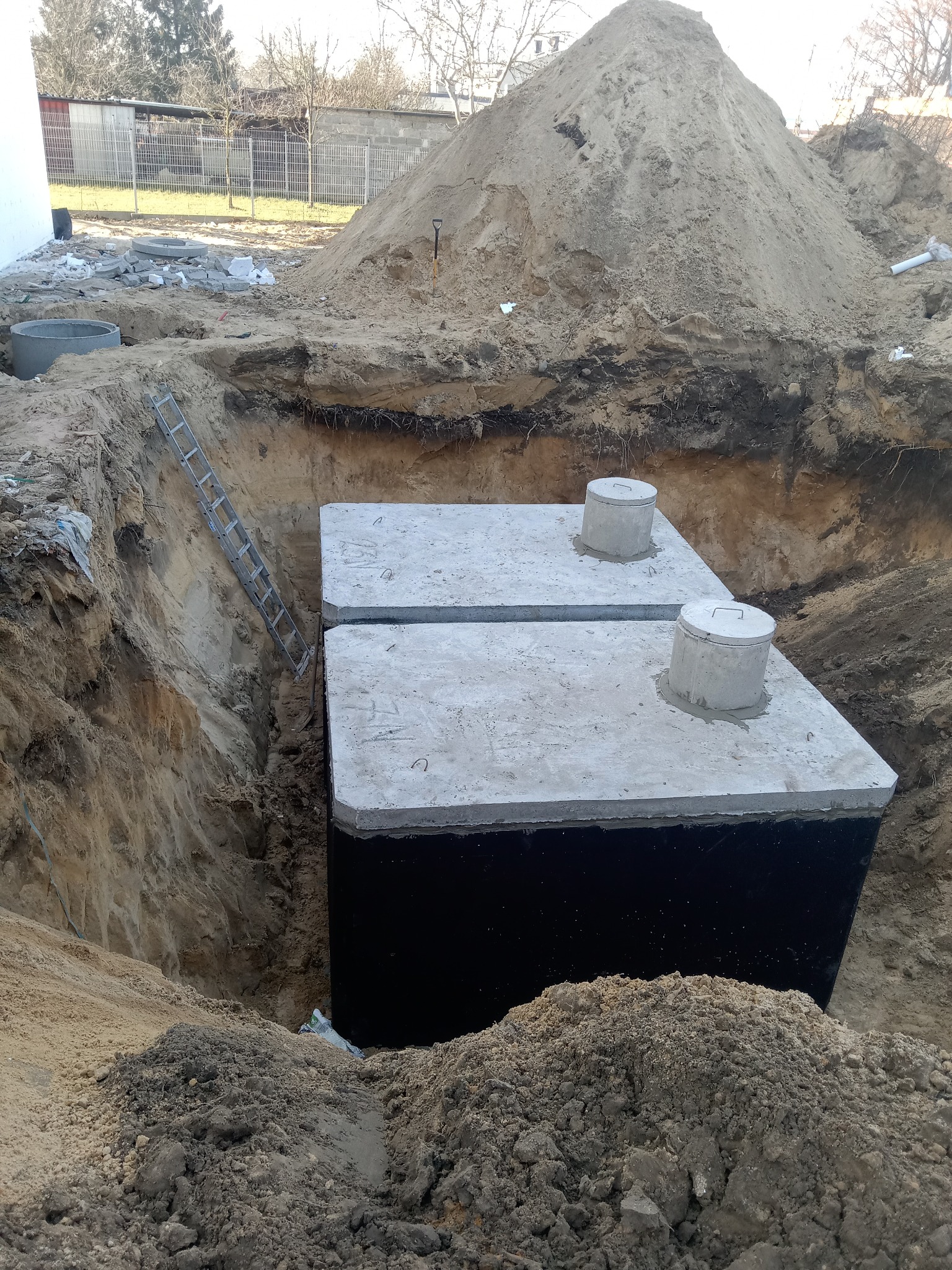 Dwa czarne, betonowe zbiorniki z pokrywami w wykopie, widoczna drabina oparta o ścianę wykopu, duża pryzma piasku w tle.