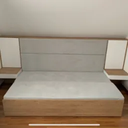 Drewniane łóżko z szarym tapicerowanym wezgłowiem i szafkami nocnymi z białymi szafkami w zabudowie w pomieszczeniu ze skosem