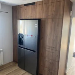 Wysoka zabudowa kuchenna z drewnopodobnymi frontami, integrująca lodówkę side-by-side z kostkarką, widoczne szuflady i szafki górne.