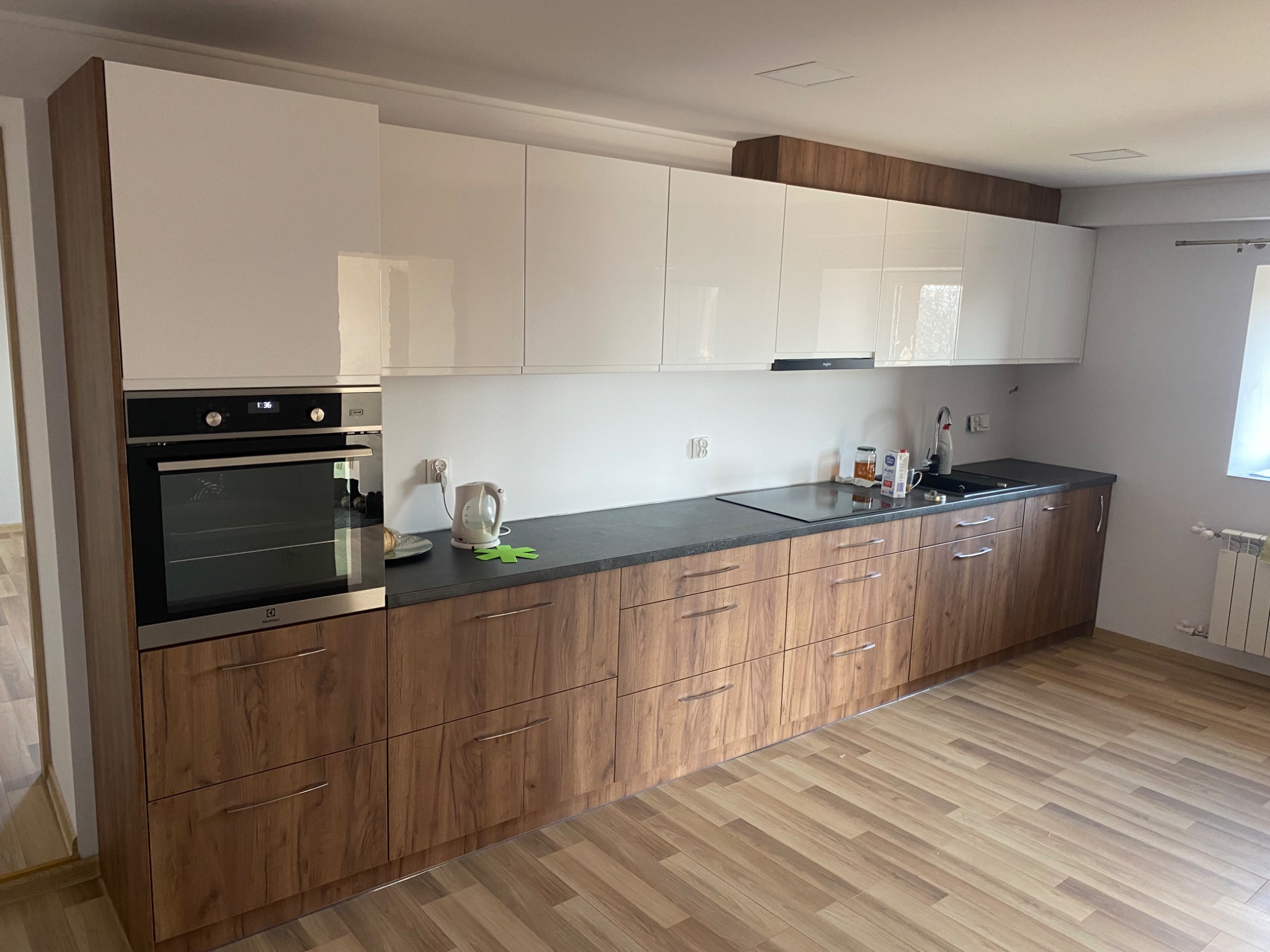 Nowoczesna kuchnia z białymi, lakierowanymi szafkami górnymi i drewnopodobnymi szafkami dolnymi z ciemnym blatem, widoczny piekarnik Electrolux i czajnik elektryczny.