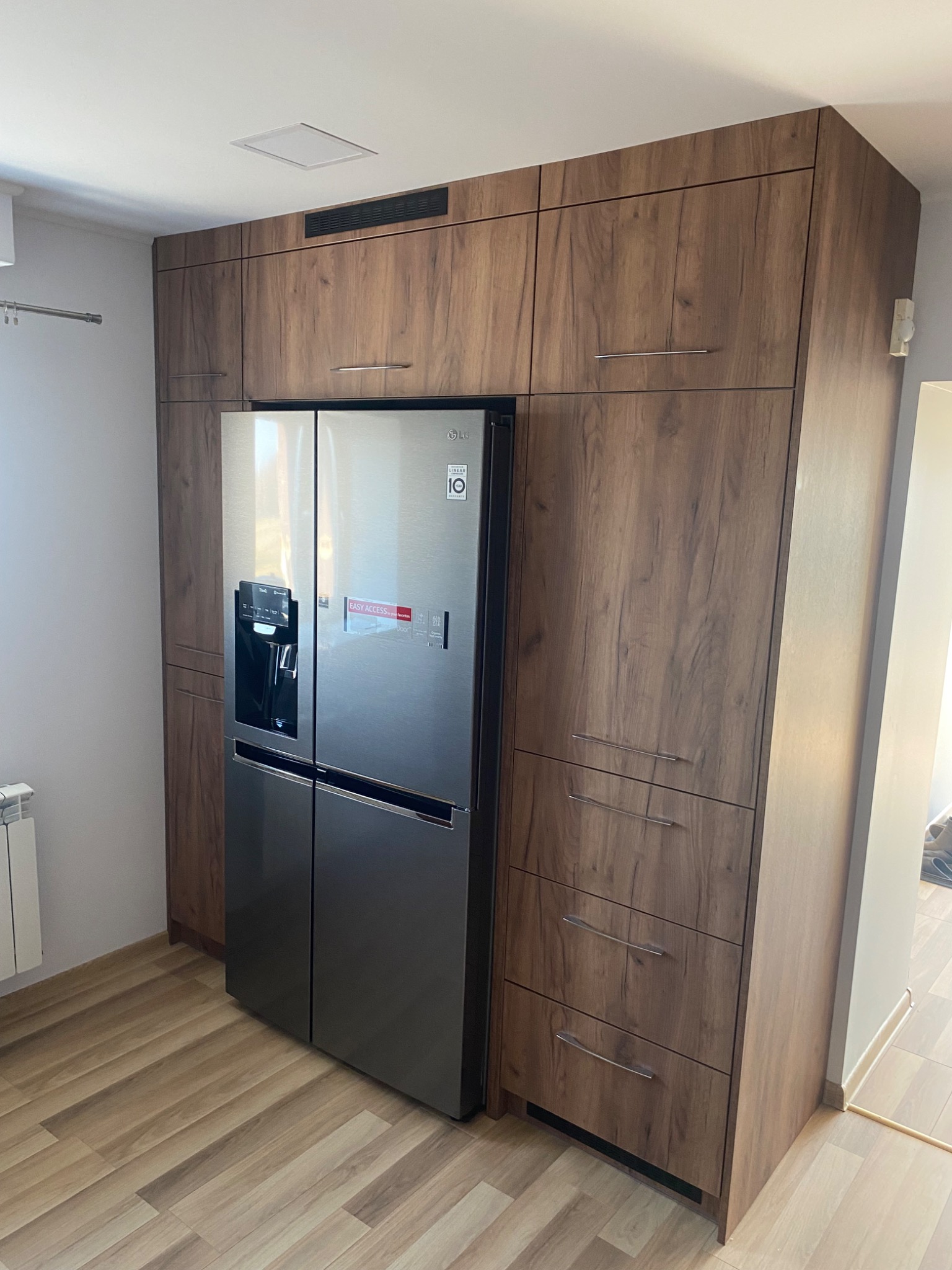 Wysoka zabudowa kuchenna z drewnopodobnymi frontami, integrująca lodówkę side-by-side z kostkarką, widoczne szuflady i szafki górne.