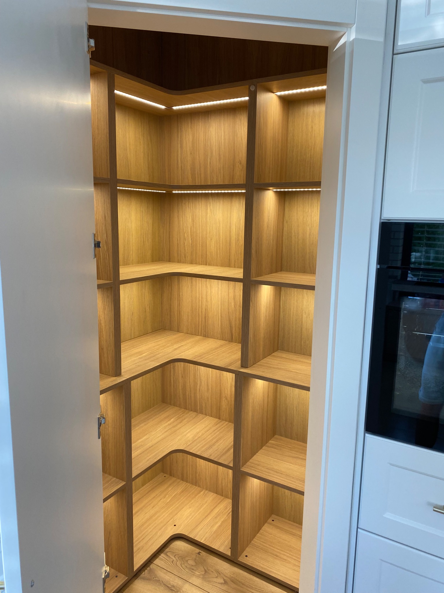 Otwarta, narożna spiżarnia z jasnego drewna z oświetleniem LED na półkach, widoczna z boku, z białą zabudową kuchenną w tle.