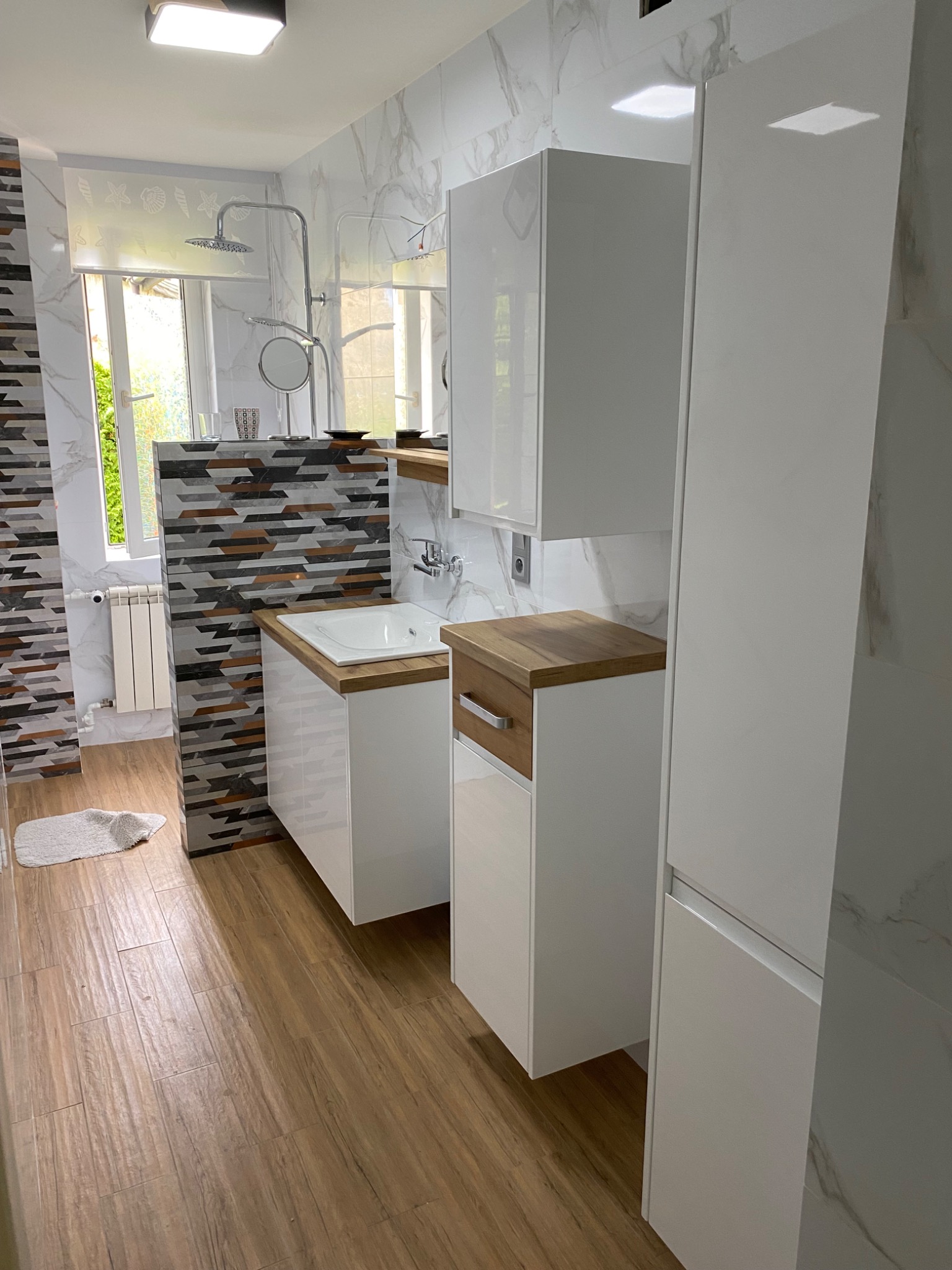 Nowoczesna łazienka z białymi szafkami na wymiar, drewnianymi blatami i panelami ściennymi imitującymi marmur oraz mozaiką, widok na kabinę prysznicową i okno.