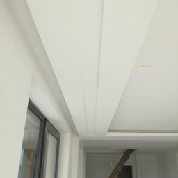 Regimex licencjonowany wykonawca firmy RIGIPS Polska. - Biały sufit z dekoracyjnymi, podłużnymi wgłębieniami i oświetleniem w nowym wnętrzu, widok z dołu, drabina w tle.