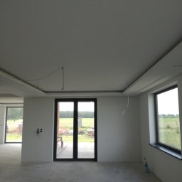 Regimex licencjonowany wykonawca firmy RIGIPS Polska. - Wnętrze w stanie deweloperskim z wykończonym sufitem podwieszanym, widoczne kable wystające z sufitu, duże okna z widokiem na zewnątrz, betonowa podłoga i butelka wody w rogu.