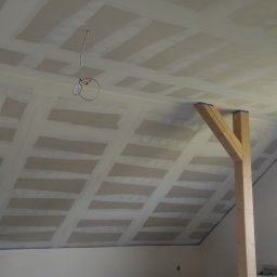 Regimex licencjonowany wykonawca firmy RIGIPS Polska. - Poddasze w trakcie remontu, widoczne płyty na skosach dachu, drewniane podpory i przewody elektryczne zwisające z sufitu, ściany przygotowane do malowania.