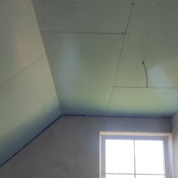 Regimex licencjonowany wykonawca firmy RIGIPS Polska. - Montaż płyt gipsowo-kartonowych na poddaszu, widoczne łączenia i wkręty, surowa ściana z oknem w tle.