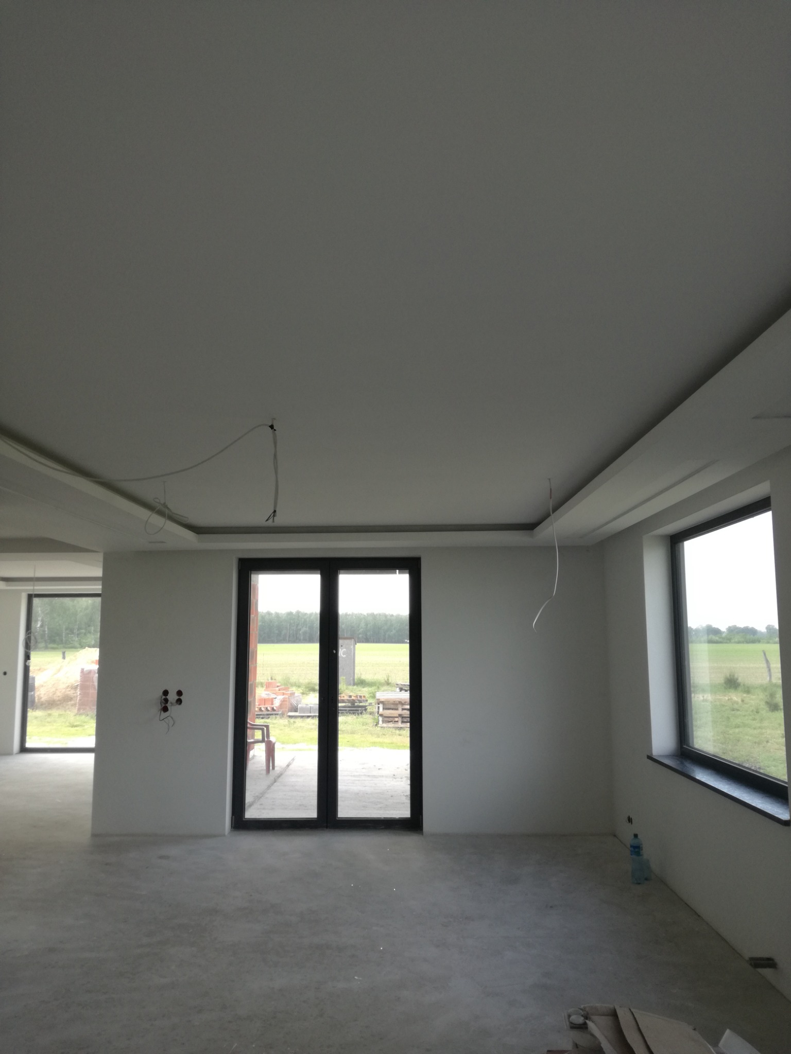 Wnętrze w stanie deweloperskim z wykończonym sufitem podwieszanym, widoczne kable wystające z sufitu, duże okna z widokiem na zewnątrz, betonowa podłoga i butelka wody w rogu.