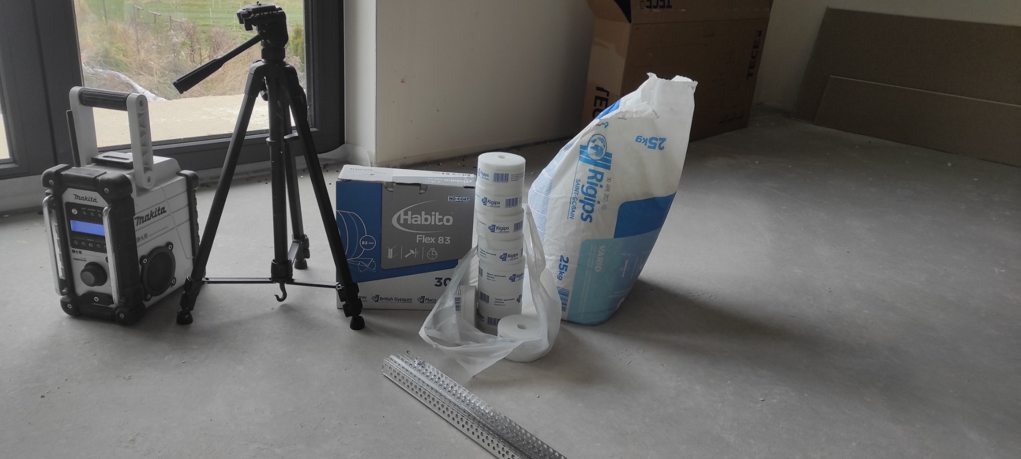 Przygotowanie do prac wykończeniowych: torba z gipsem szpachlowym, rolki taśmy, profil aluminiowy, pudełko płyty Habito Flex, radio budowlane Makita i statyw na podłodze w surowym wnętrzu.
