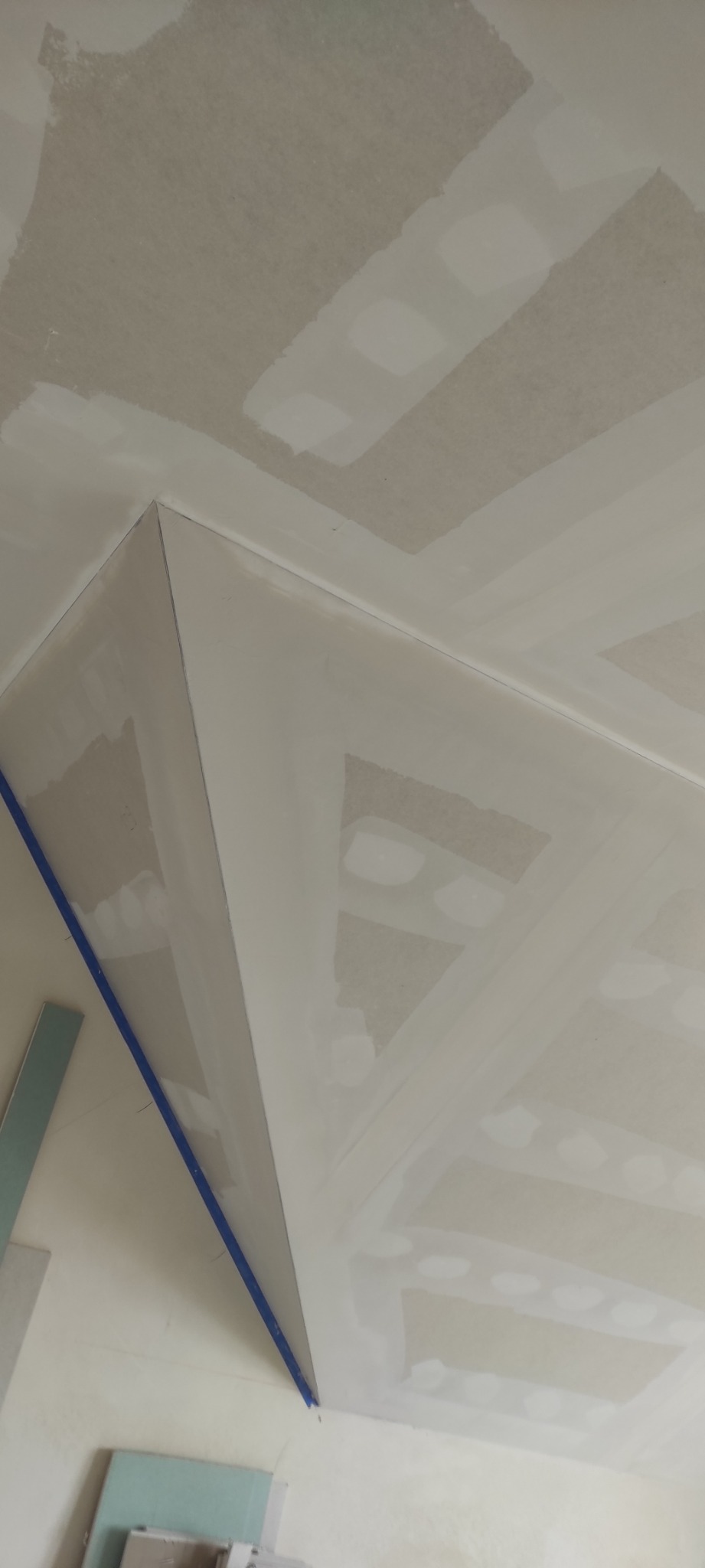 Sufit z płyt kartonowo-gipsowych w trakcie szpachlowania, widoczne łączenia płyt z taśmą i szpachlą, fragment ściany z niebieską taśmą malarską.