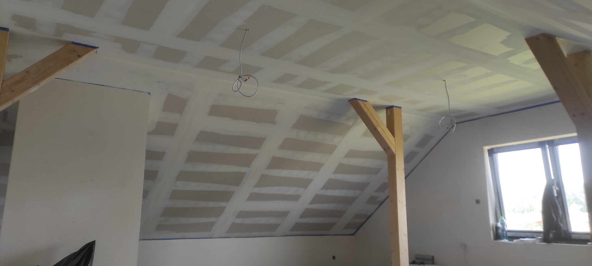 Poddasze w trakcie remontu, widoczne płyty na skosach dachu, drewniane podpory i przewody elektryczne zwisające z sufitu, ściany przygotowane do malowania.