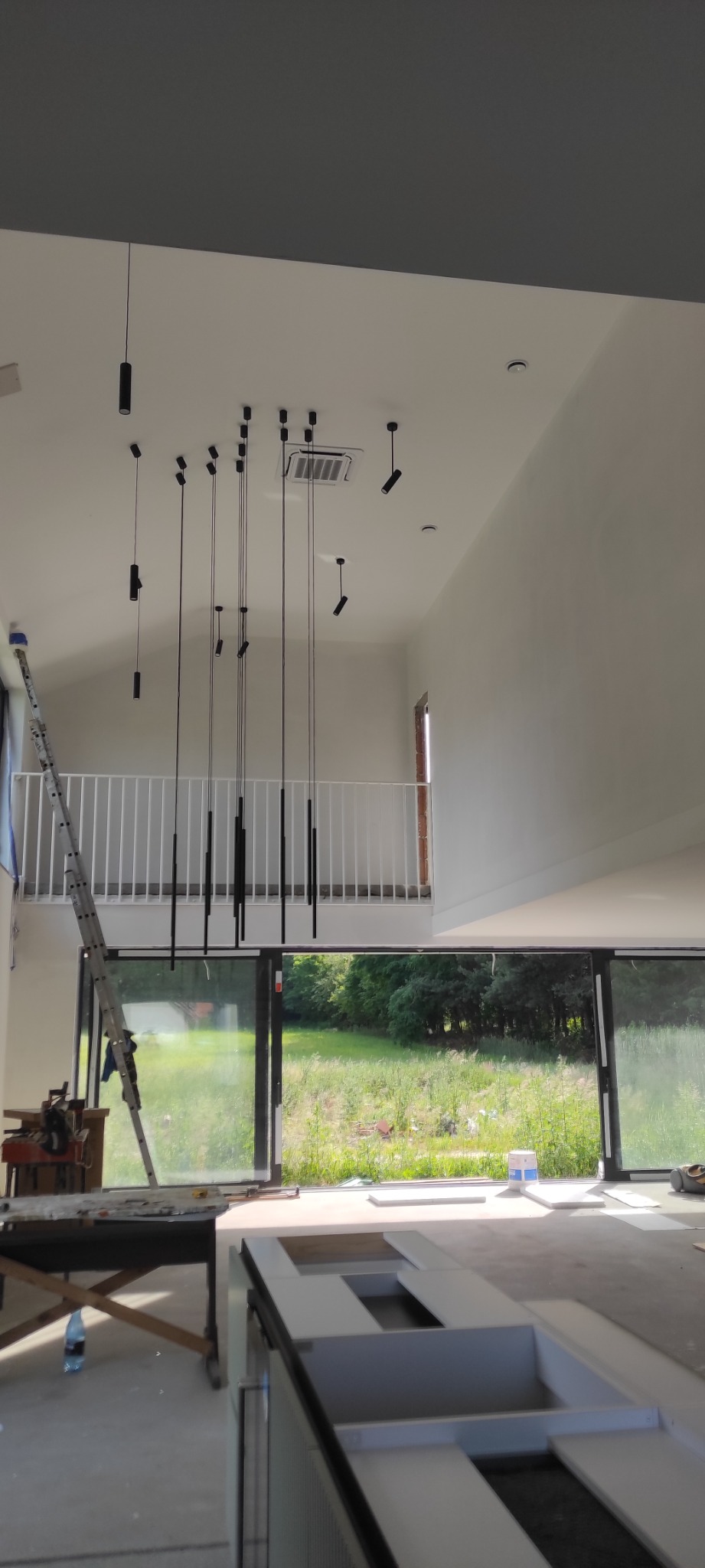 Wnętrze w trakcie wykańczania z wysokim sufitem i zainstalowanym oświetleniem punktowym, widoczna drabina i elementy zabudowy kuchennej.