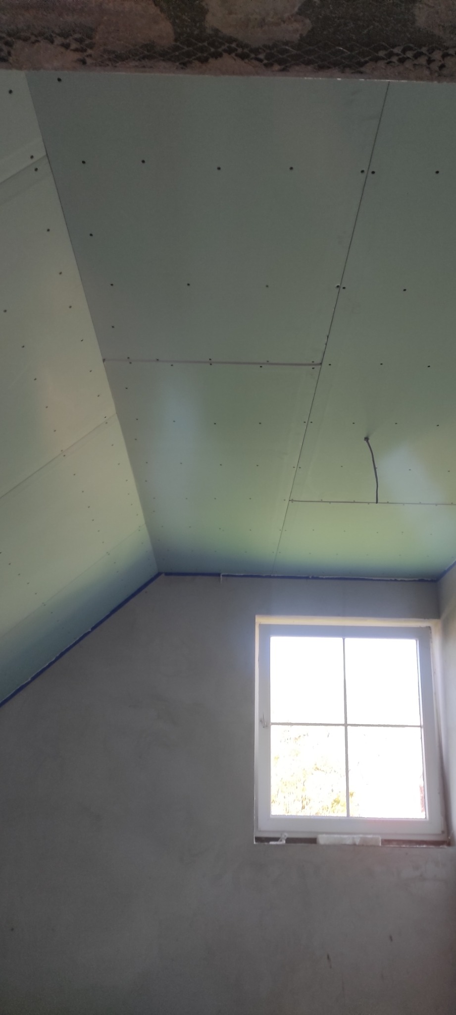 Montaż płyt gipsowo-kartonowych na poddaszu, widoczne łączenia i wkręty, surowa ściana z oknem w tle.