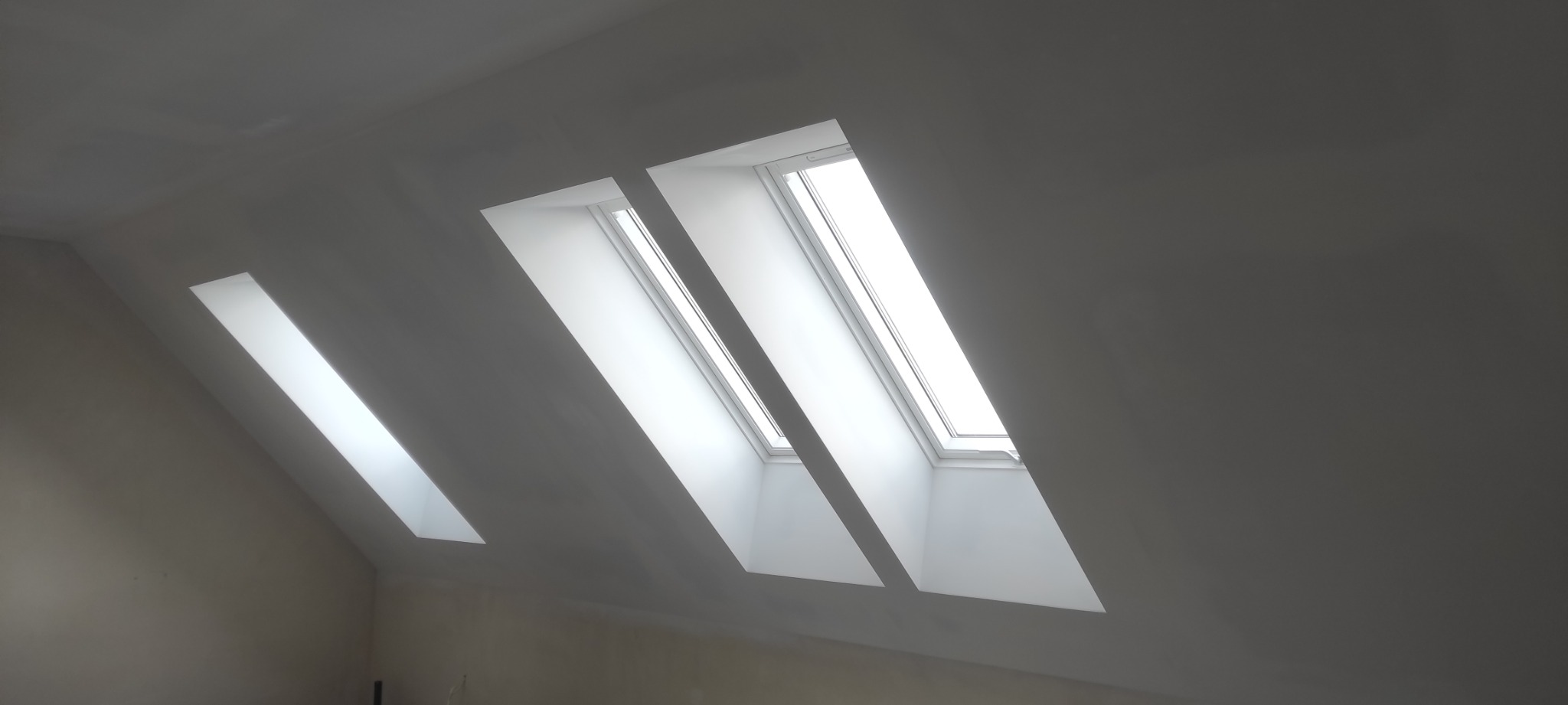 Poddasze z trzema oknami dachowymi po szpachlowaniu i malowaniu na biało, widoczne nierówności na ścianie bocznej.