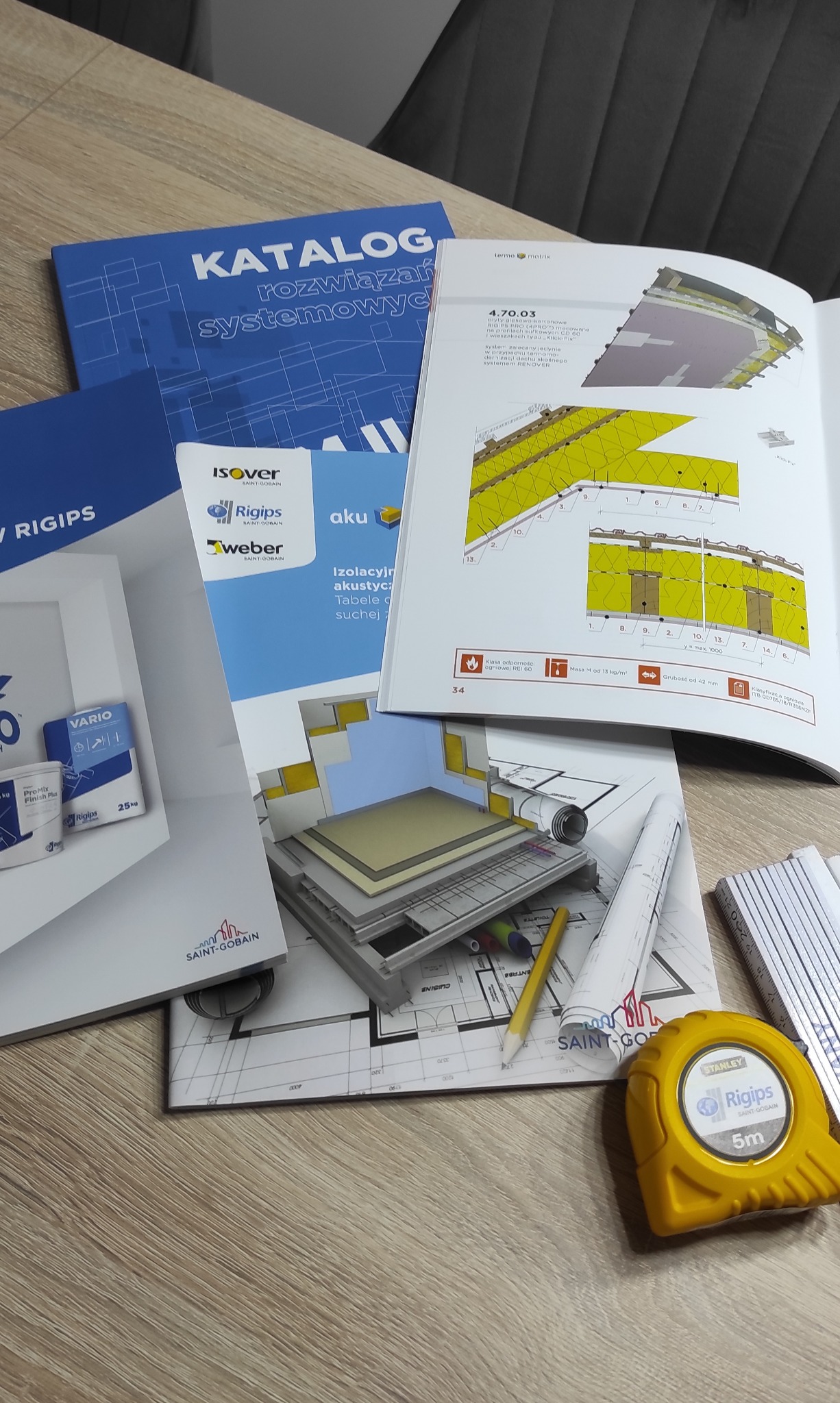 Zestaw materiałów informacyjnych i narzędzi budowlanych na drewnianym blacie: katalogi rozwiązań systemowych, broszury Rigips, Isover i Weber, zwinięte plany, ołówek i żółta miara zwijana Stanley...