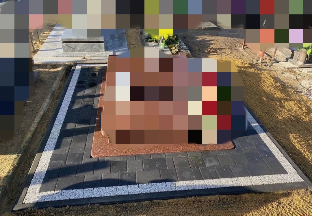 Ciemnoszara kostka brukowa z białym obramowaniem wokół nagrobka z czerwonego granitu.