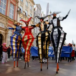 Cztery osoby na szczudłach w kostiumach błaznów, prezentujące się na tle kamienic w Gdańsku, obserwowane przez tłum widzów.