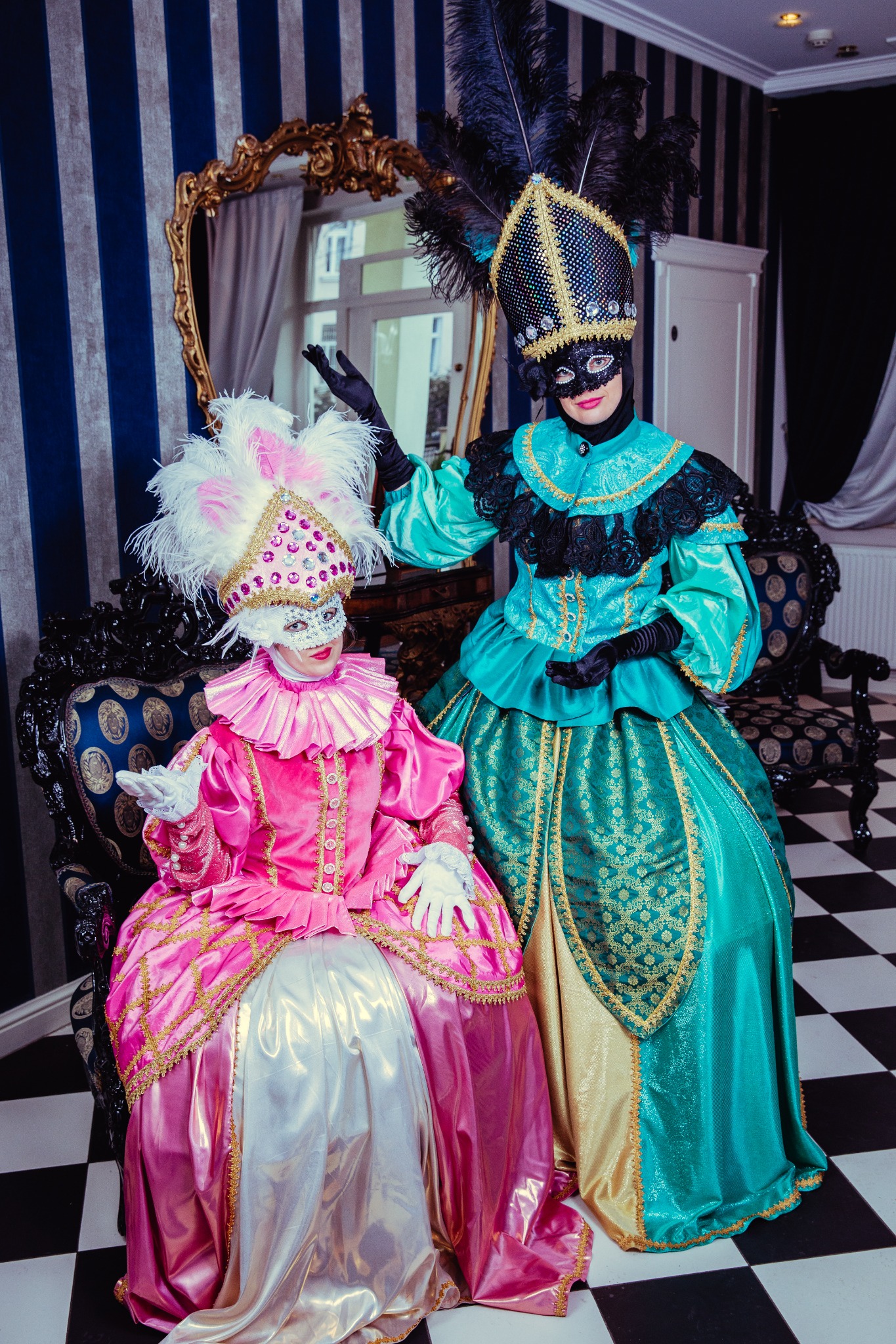 Dwie osoby w bogato zdobionych, kostiumach weneckich z piórami i maskami, siedzące na ozdobnych krzesłach na tle pokoju z tapetą w paski i szachownicą na podłodze.