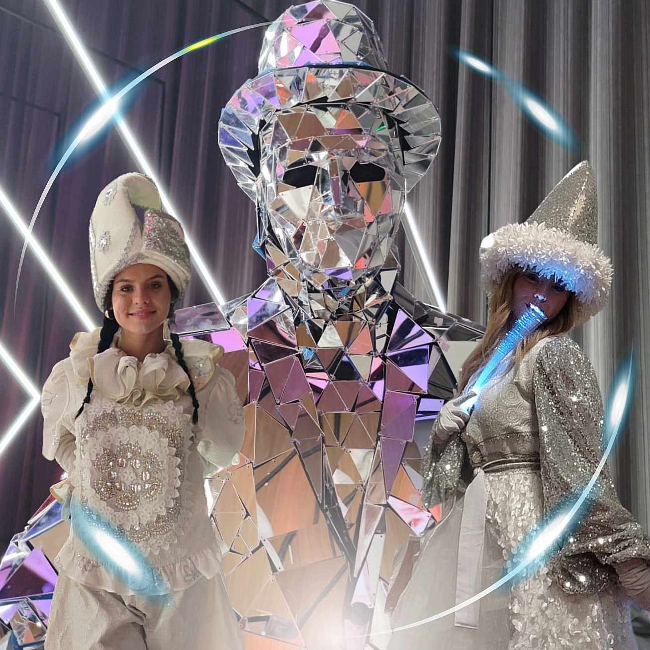 Dwie uśmiechnięte kobiety w błyszczących kostiumach obok postaci w lustrzanym kostiumie z kapeluszem, na tle ciemnej zasłony i neonowych świateł.