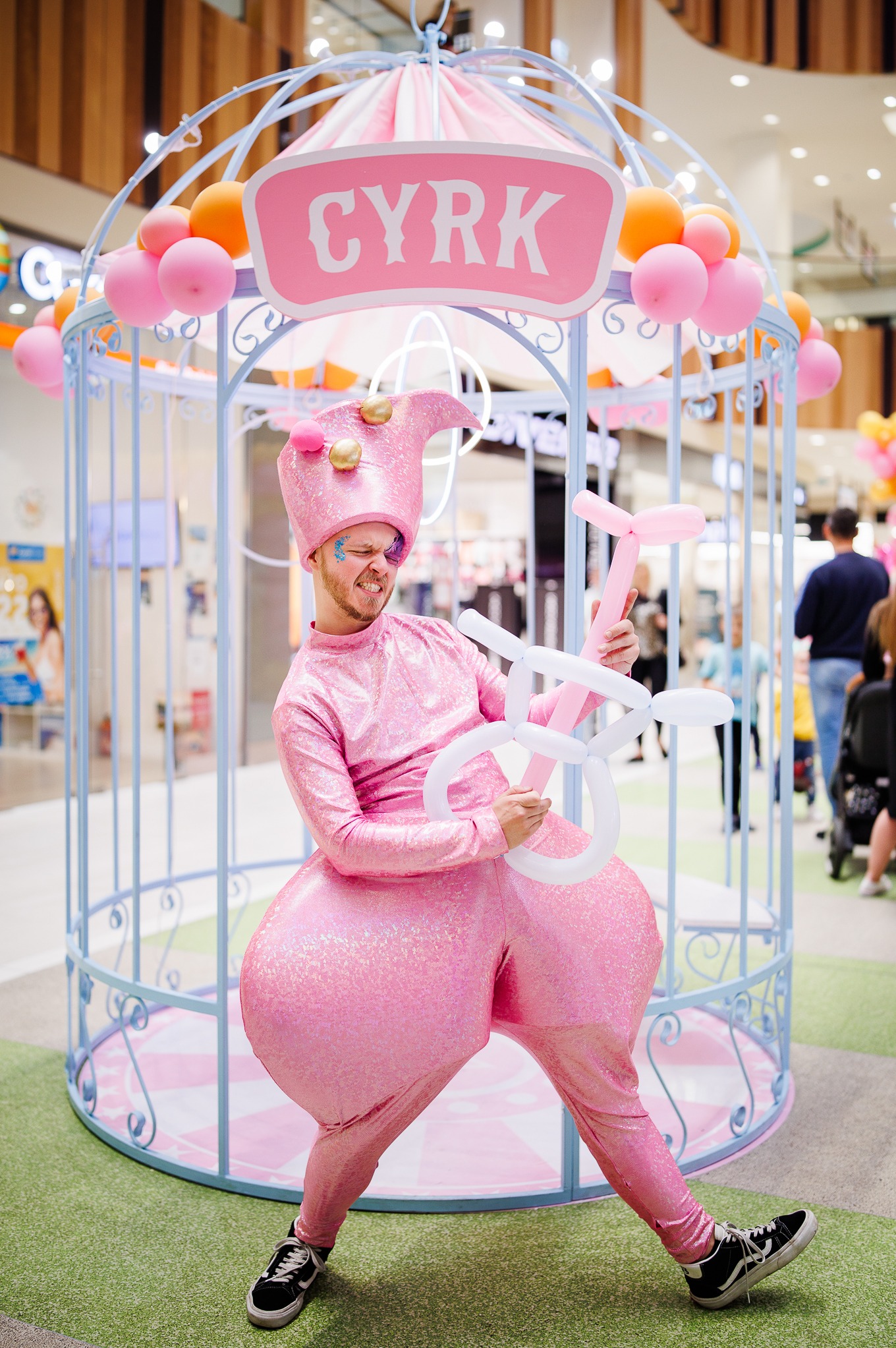 Mężczyzna w błyszczącym, różowym stroju cyrkowym i czapce, z brokatem pod okiem, modeluje balonowe zwierzę na tle dekoracji z napisem 'CYRK' i balonami.