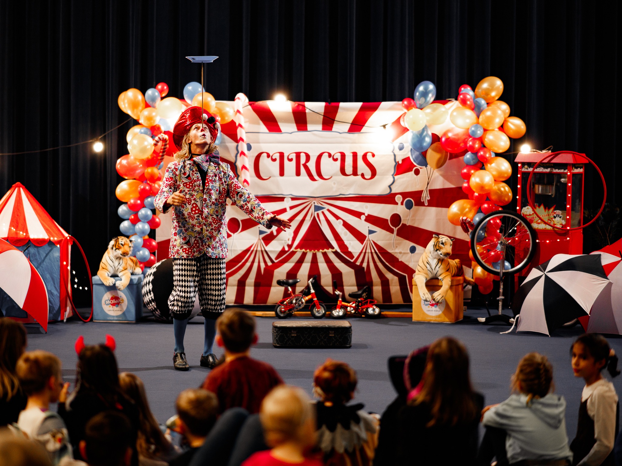 Artysta cyrkowy w kolorowym stroju balansuje talerzem na głowie podczas występu dla dzieci, na scenie dekoracje cyrkowe, balony i pluszowe tygrysy.