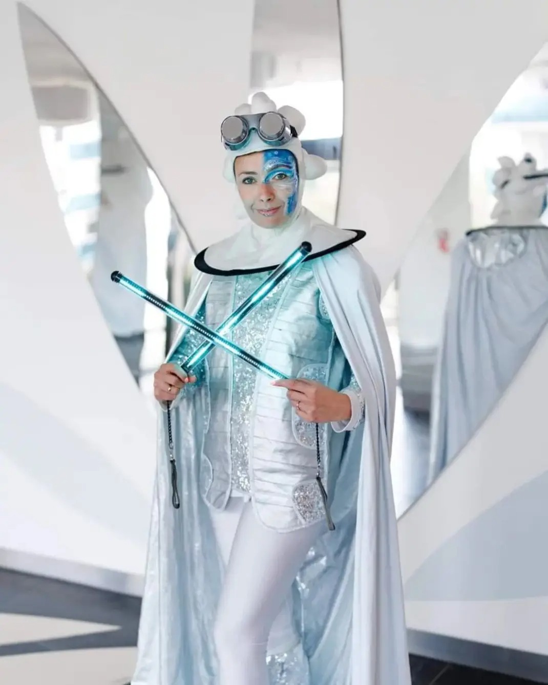 Animator w srebrnym kostiumie z futurystycznym makijażem i świetlistymi pałeczkami skrzyżowanymi na tle białych paneli.