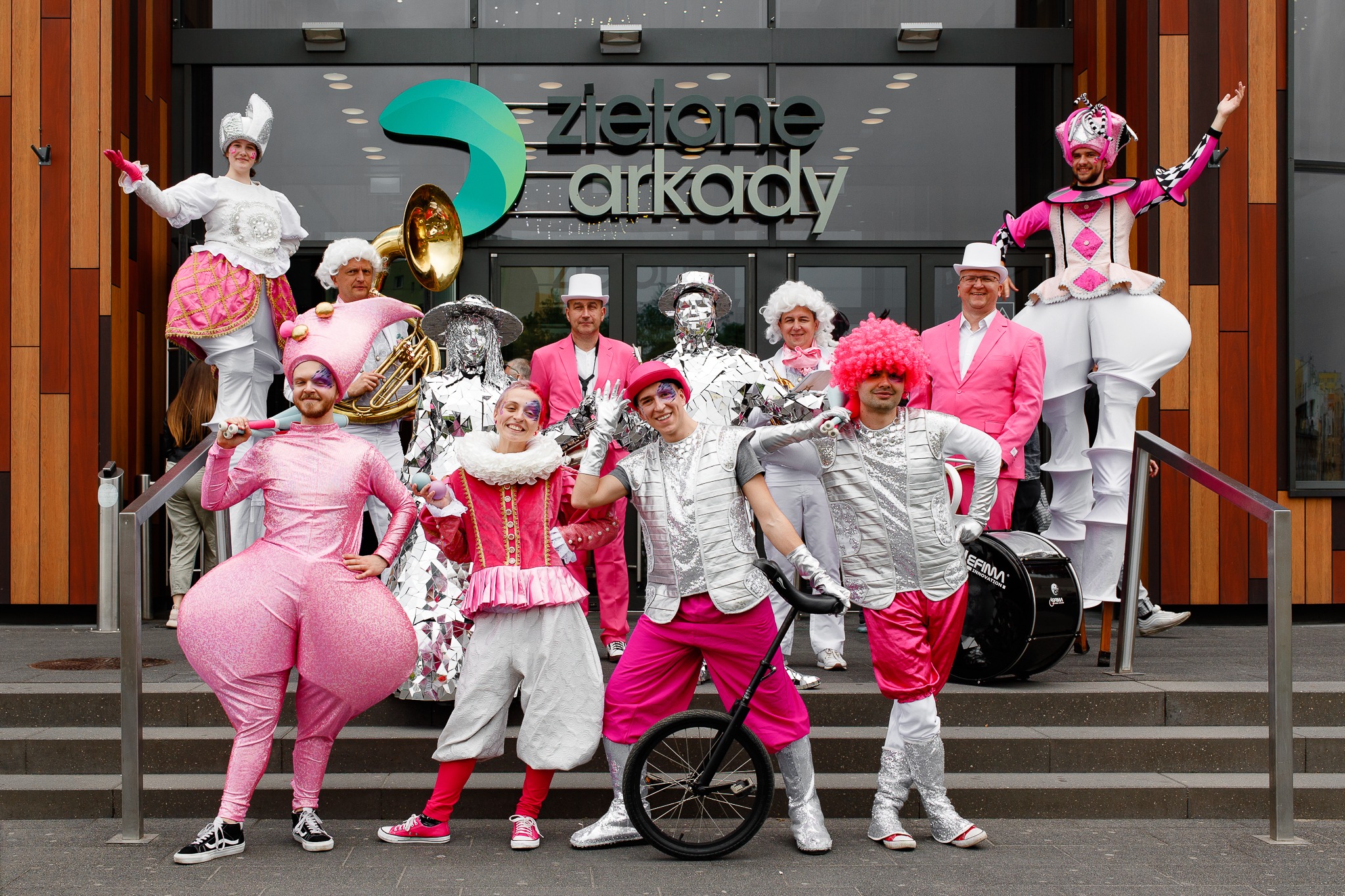 Grupa performerów w kostiumach, w tym osoba na szczudłach i z monocyklem, pozuje na schodach przed budynkiem z napisem 'Zielone Arkady'.
