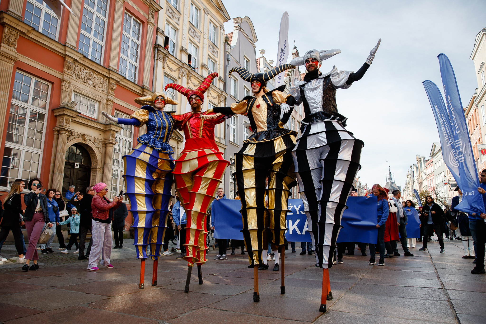 Cztery osoby na szczudłach w kostiumach błaznów, prezentujące się na tle kamienic w Gdańsku, obserwowane przez tłum widzów.