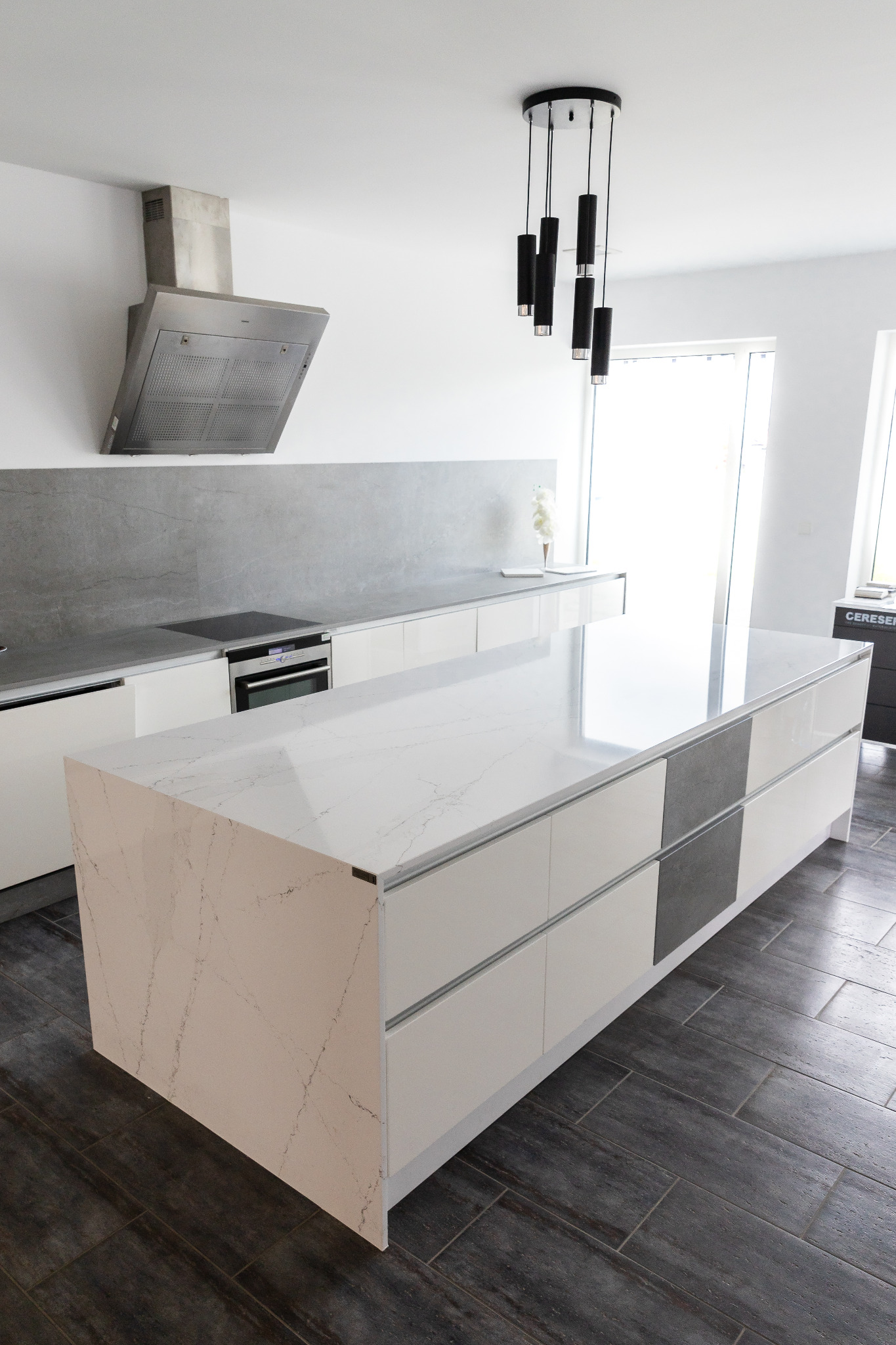 Nowoczesna wyspa kuchenna z blatem o wzorze marmuru, połączona z białymi szafkami i szarymi akcentami, uzupełniona o designerski okap i lampę wiszącą.