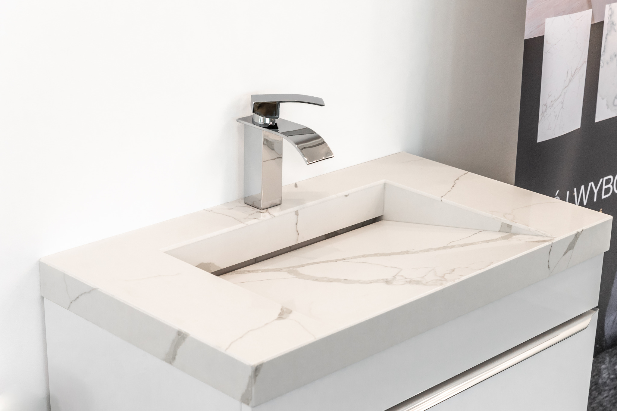 Biała umywalka wpuszczona w blat z konglomeratu z szarymi żyłkami, minimalistyczna bateria umywalkowa w tle biała ściana i fragment ekspozycji materiałów wykończeniowych.