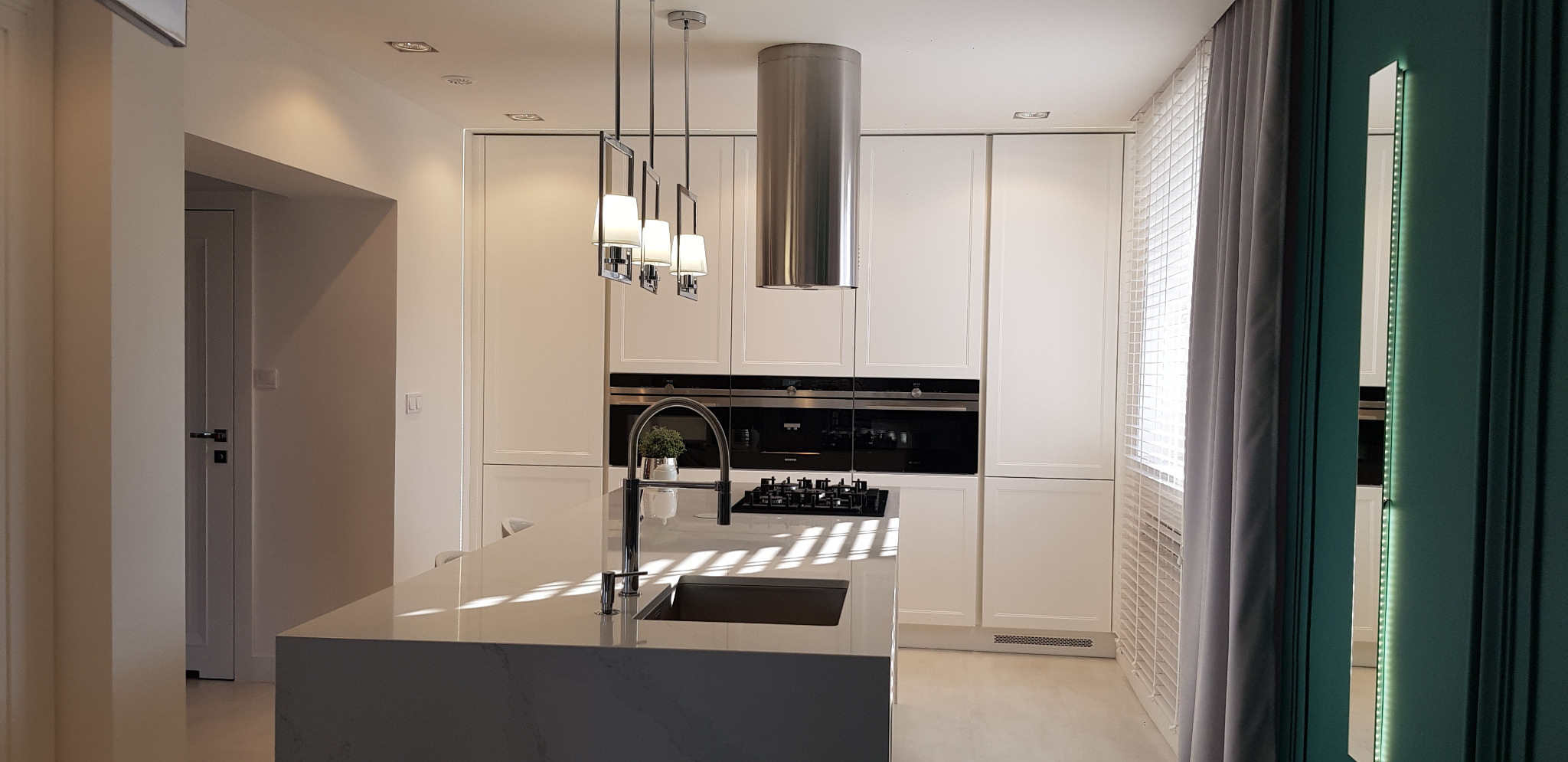 Nowoczesna, jasna kuchnia z wyspą i blatem z jasnego kamienia, oświetlona wiszącymi lampami. Na blacie widoczny zlewozmywak i bateria w kolorze czarnym oraz roślina doniczkowa. W tle zabudowa...