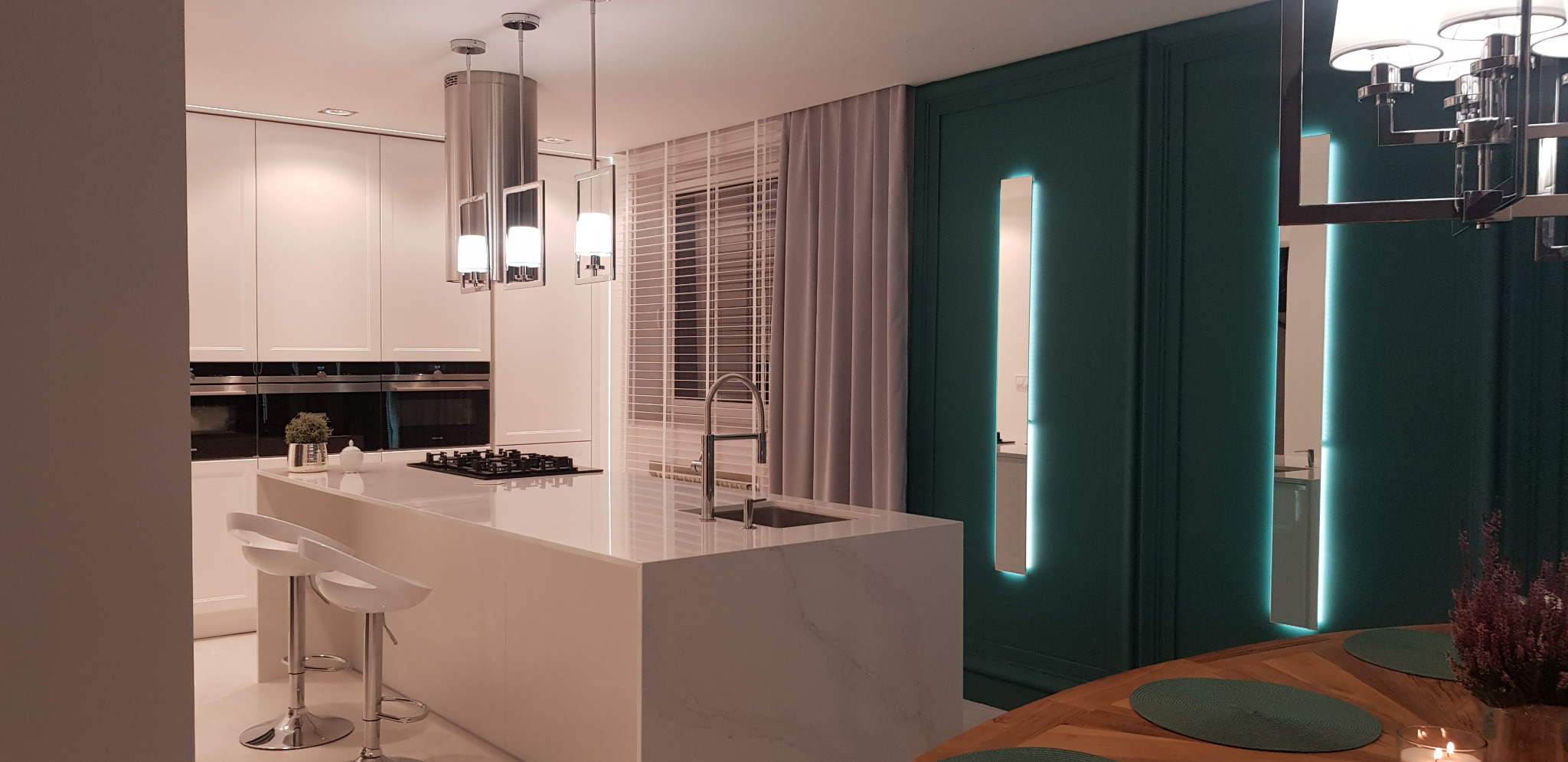 Nowoczesna kuchnia z białym blatem kamiennym na wyspie, srebrnymi hokerami i oświetleniem LED w zielonej ścianie, widok na okno z żaluzjami.