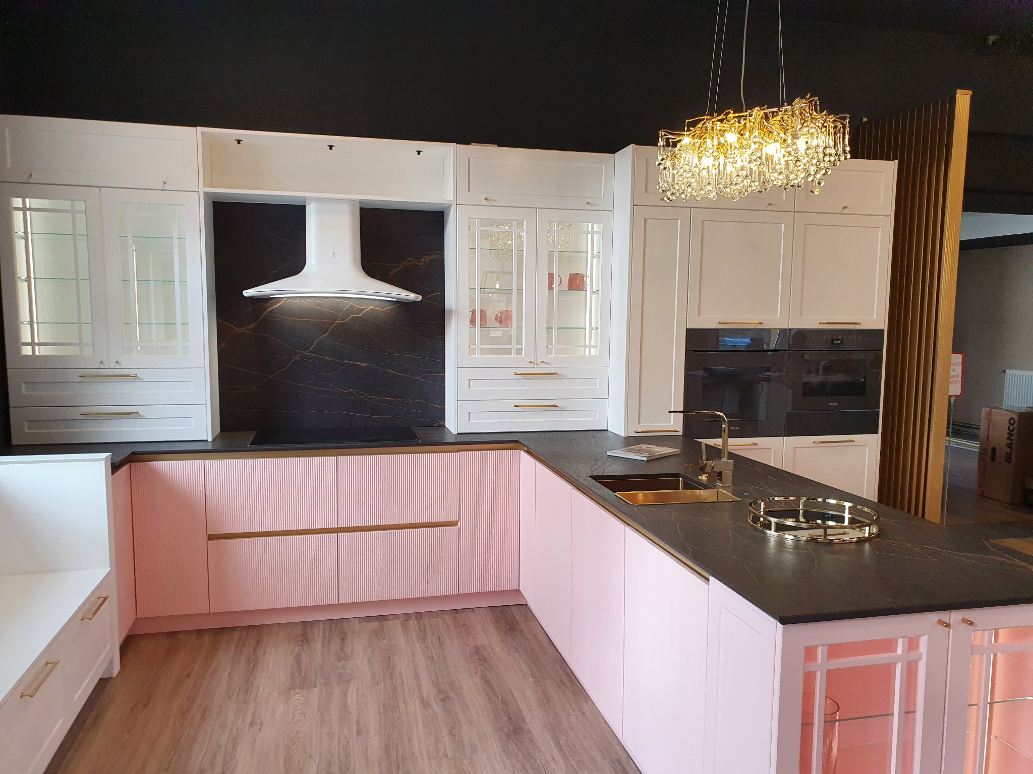 Nowoczesna kuchnia z różowymi szafkami dolnymi z ryflowanym frontem, białymi szafkami górnymi z przeszkleniami, czarnym blatem z wyraźnym użyleniem i złotymi detalami, oświetlona dekoracyjnym...