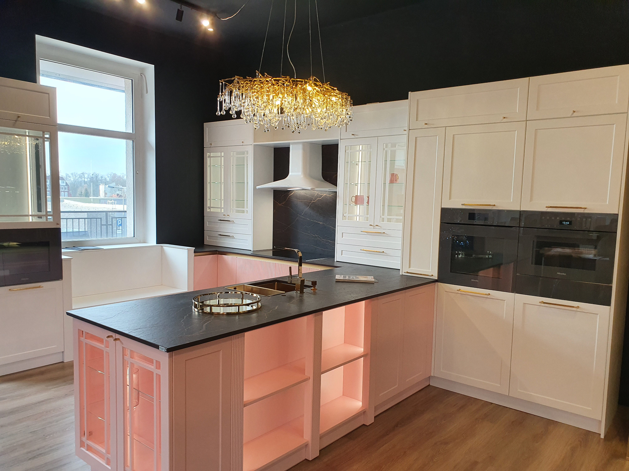 Nowoczesna kuchnia z wyspą o blacie z ciemnego kamienia, różowe szafki z podświetleniem LED, białe szafki wiszące i piekarniki w zabudowie, złoty żyrandol.