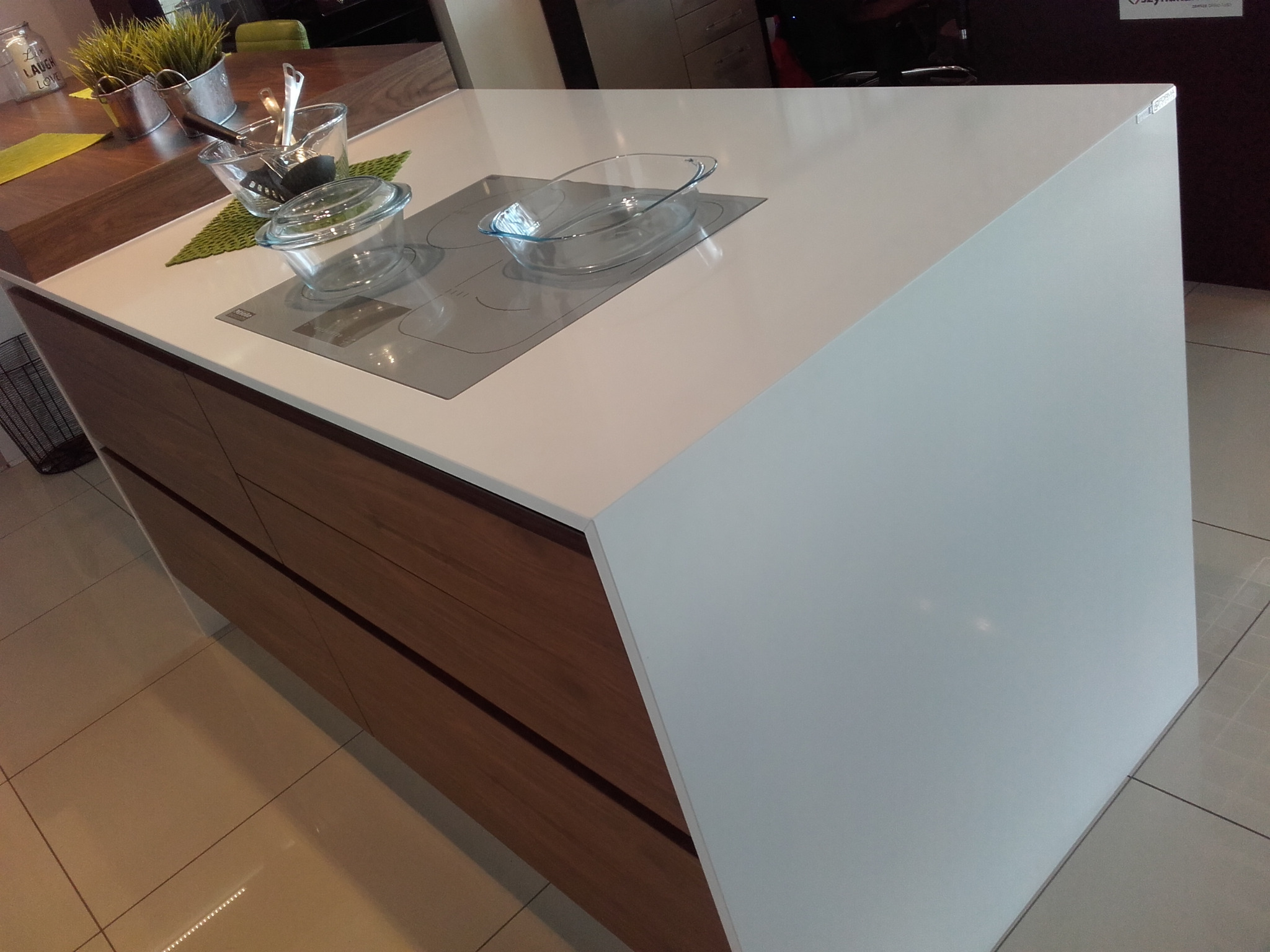 Biały blat kamienny kuchennej wyspy z wbudowaną płytą indukcyjną i szklanymi naczyniami, na drewnianej podstawie z szufladami.