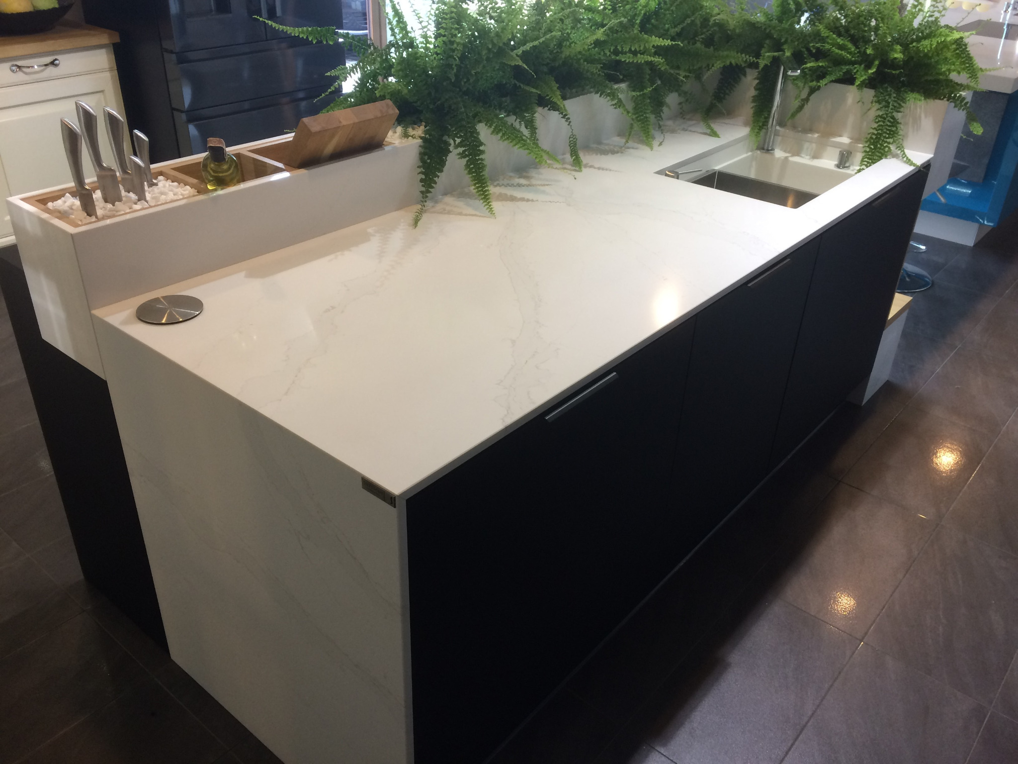 Biały blat kuchenny z szarymi żyłkami, czarne szafki, wbudowany zlew i donica z paprotkami, widoczne noże w pojemniku z białymi kamykami i butelka z olejem.