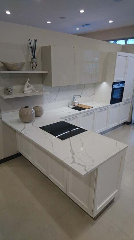Nowoczesna, jasna kuchnia z białymi szafkami i blatem z białego kamienia z wyraźnym szarym żyłkowaniem, wyposażona w płytę indukcyjną i piekarnik w zabudowie.