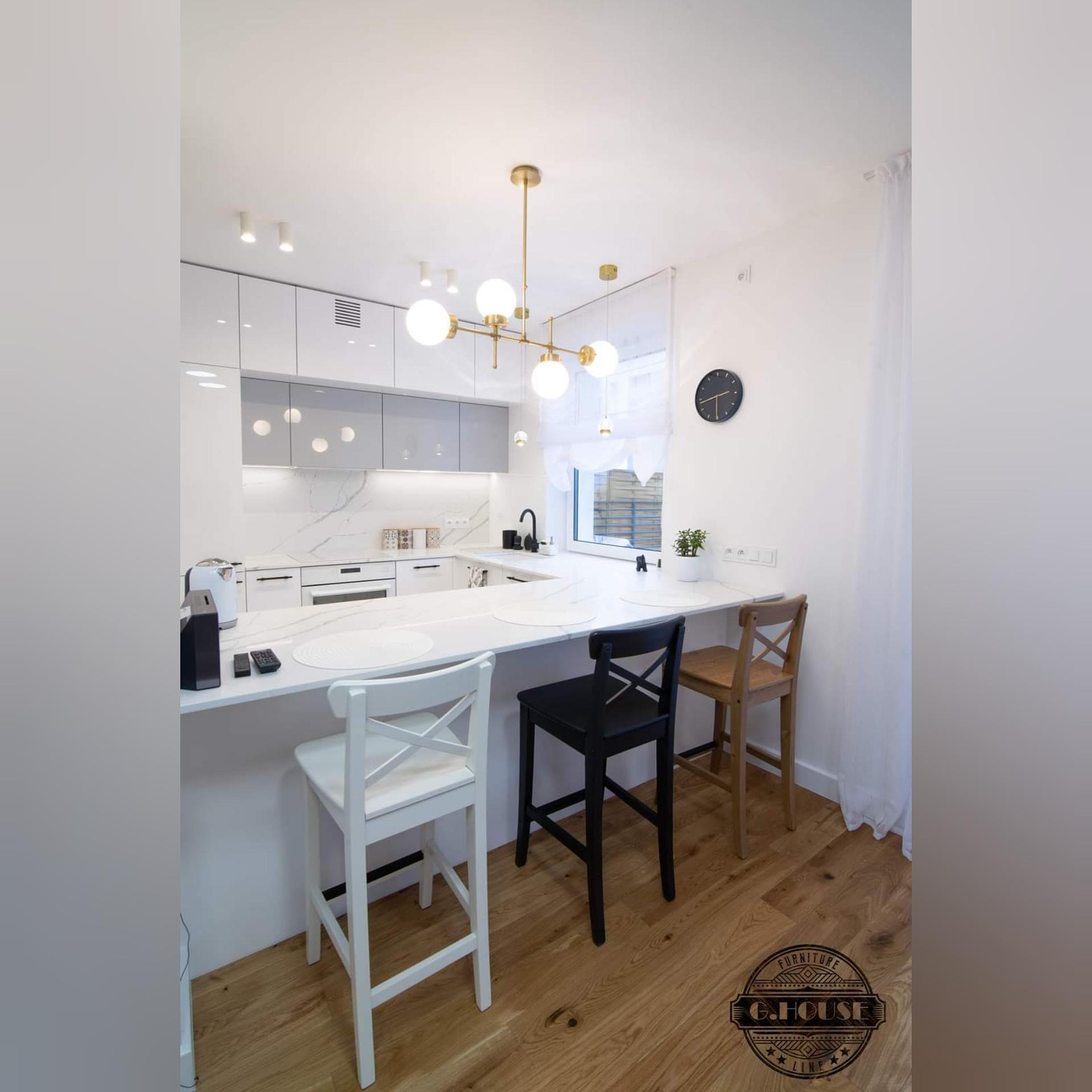 Nowoczesna kuchnia z białymi szafkami i blatem imitującym marmur, oświetlona złotą lampą wiszącą i trzema krzesłami barowymi: białym, czarnym i drewnianym. Widoczny minimalistyczny zegar na ścianie...