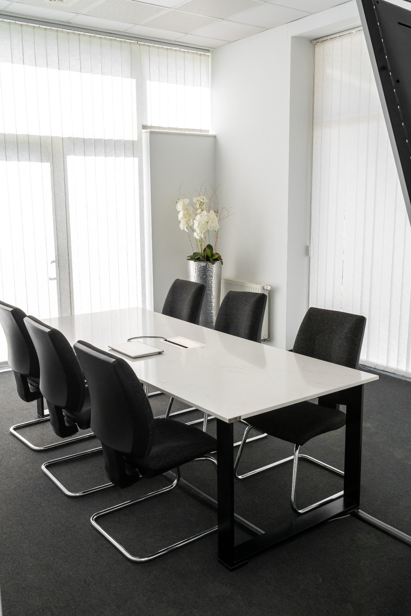 Biały stół konferencyjny z blatem o wzorze kamienia, otoczony czarnymi krzesłami na chromowanych nogach, w jasnym biurze z pionowymi żaluzjami i srebrną donicą z białymi kwiatami.
