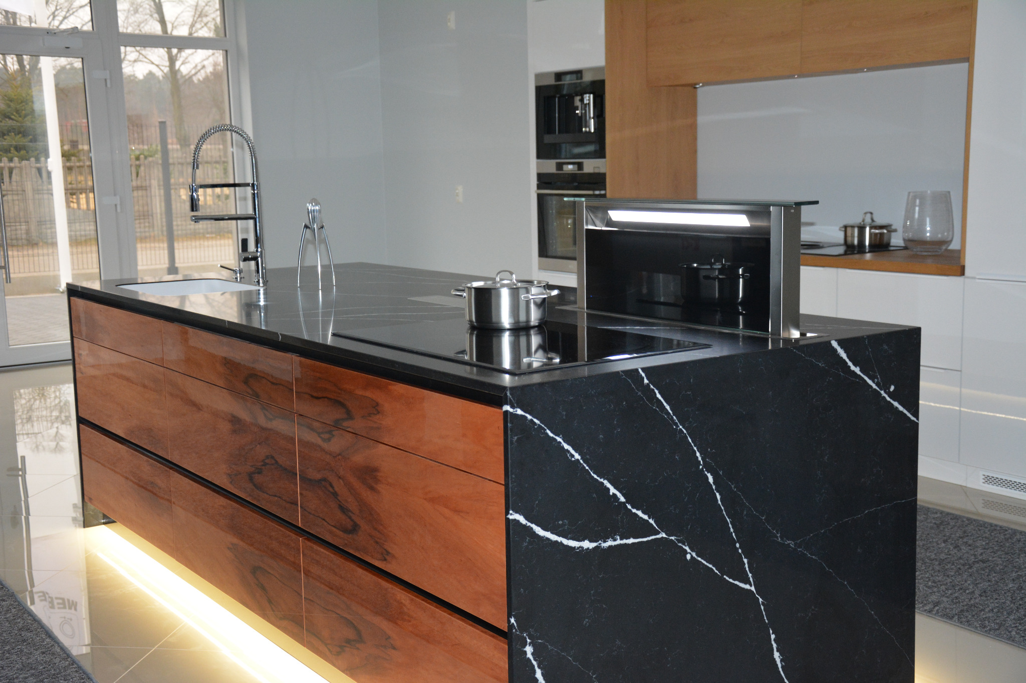 Nowoczesna wyspa kuchenna z czarnym blatem z białym żyłkowaniem, drewnianymi frontami szuflad i podświetleniem LED u dołu, uzupełniona o zlew, baterię i płytę indukcyjną z pochłaniaczem.