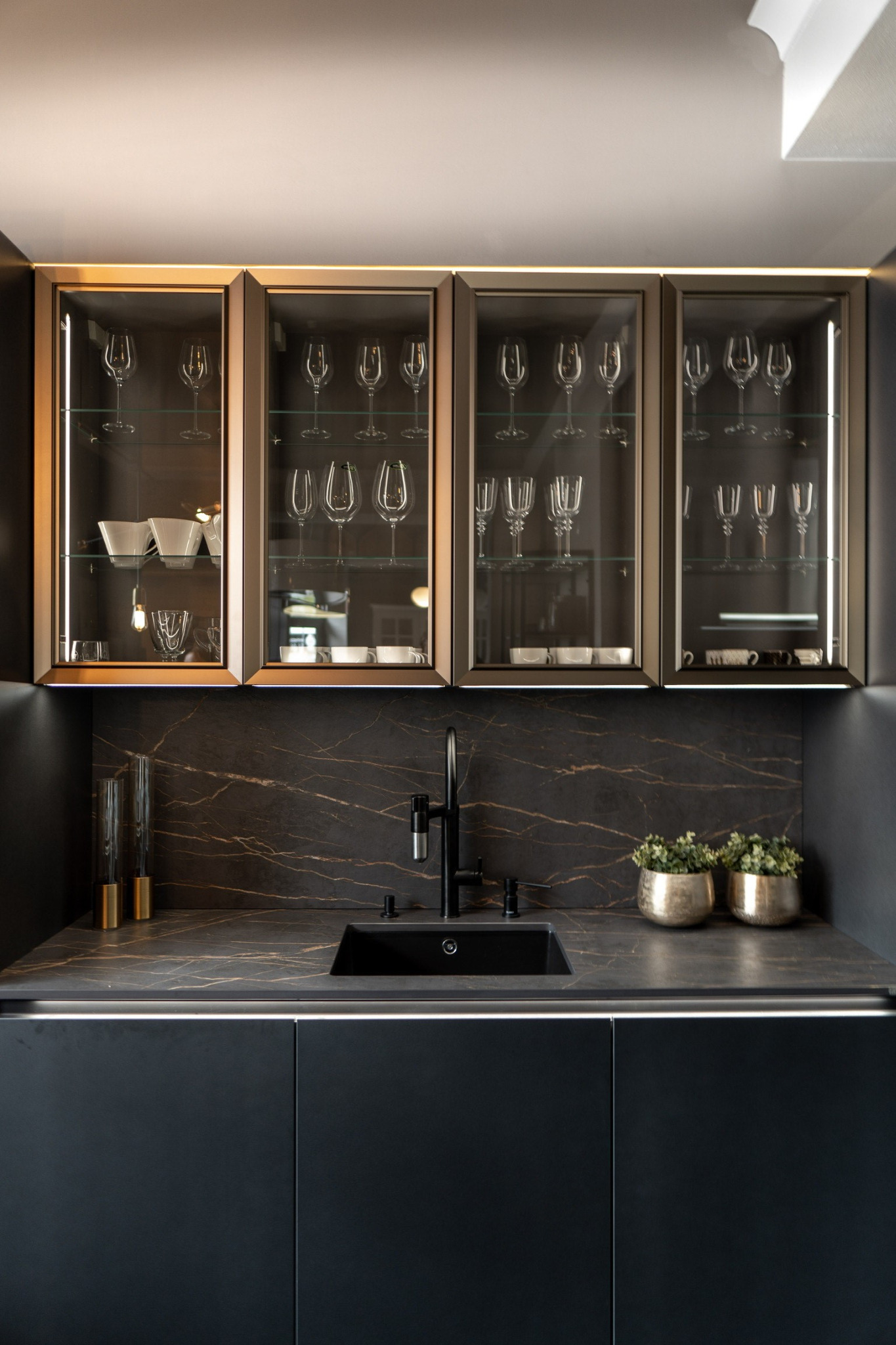 Ciemna kuchnia z blatem imitującym marmur, czarnym zlewem i złotymi akcentami, szklane szafki wiszące z oświetleniem LED eksponują kieliszki i naczynia.
